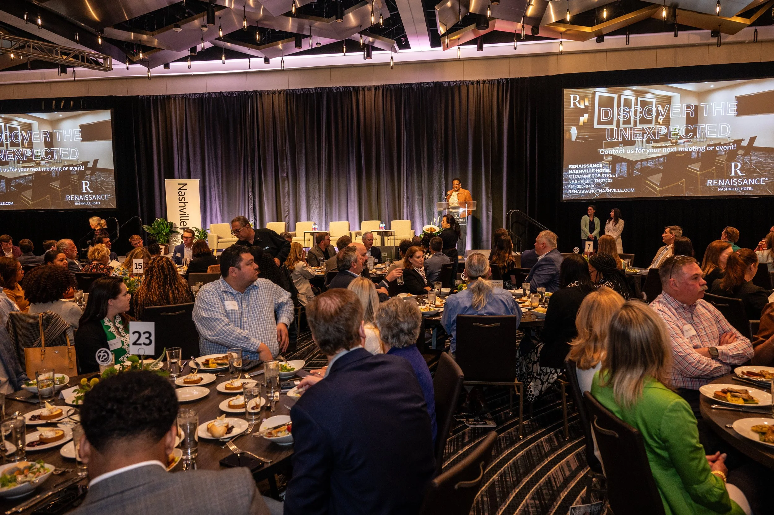 A conference or event held in a large ballroom with round tables and many attendees seated, facing a stage with a speaker at a podium. Large screens on either side of the stage display event branding and details. The room is decorated with dark curta