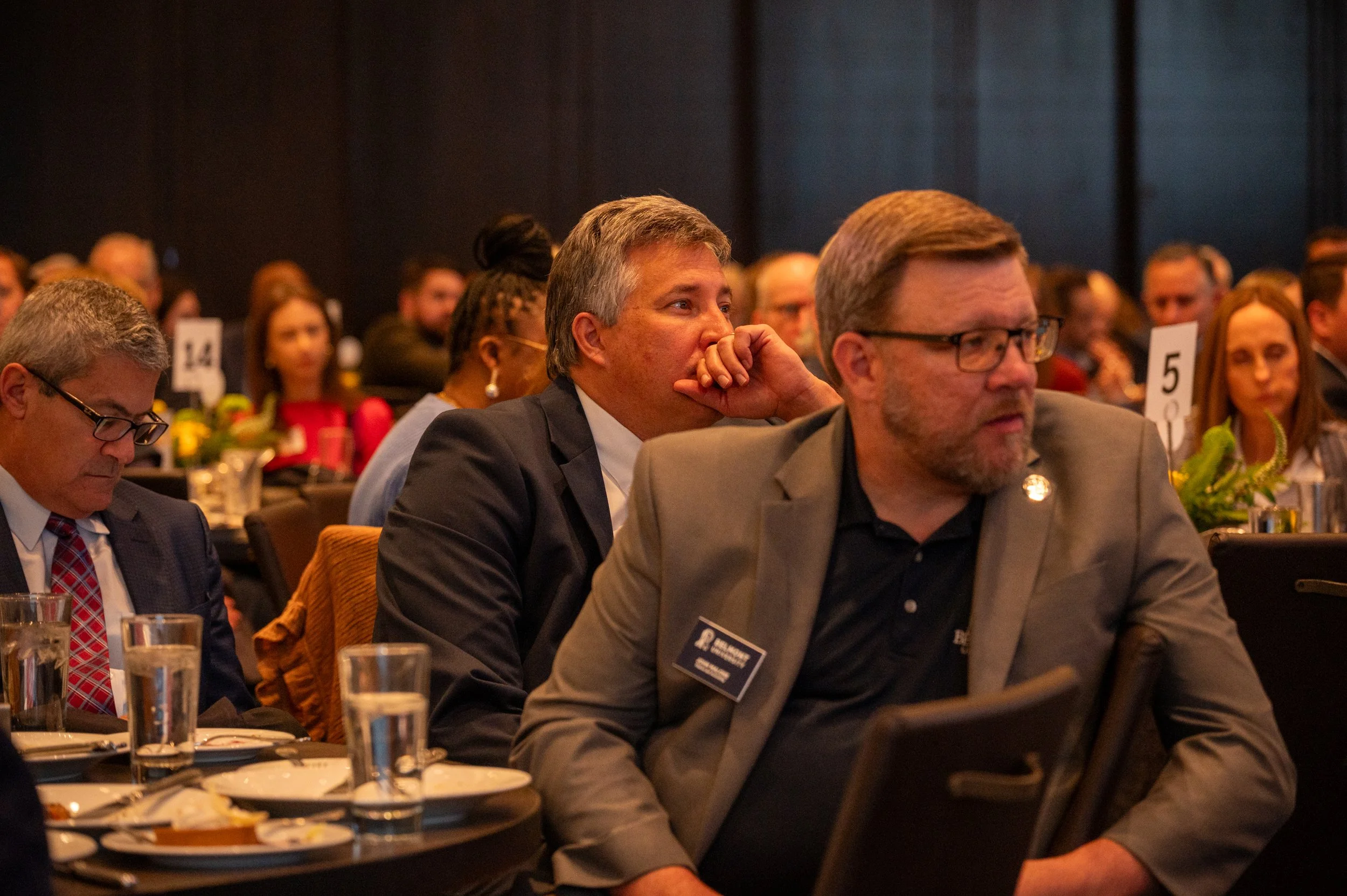 People attending a formal event or conference sitting at round tables with plates and glasses, listening attentively.