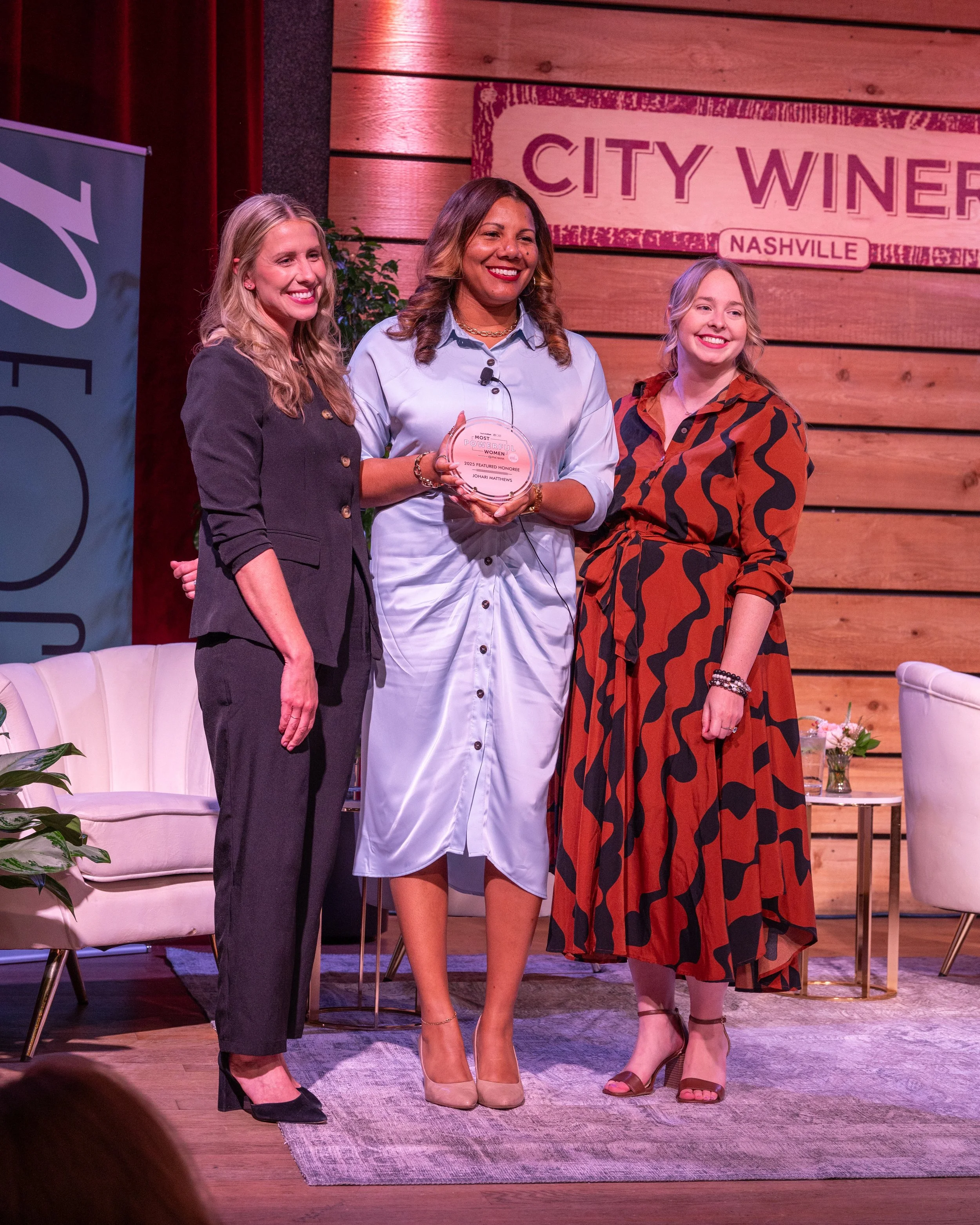 Three women standing on a stage, with one woman holding an award, at an event for women in Nashville.