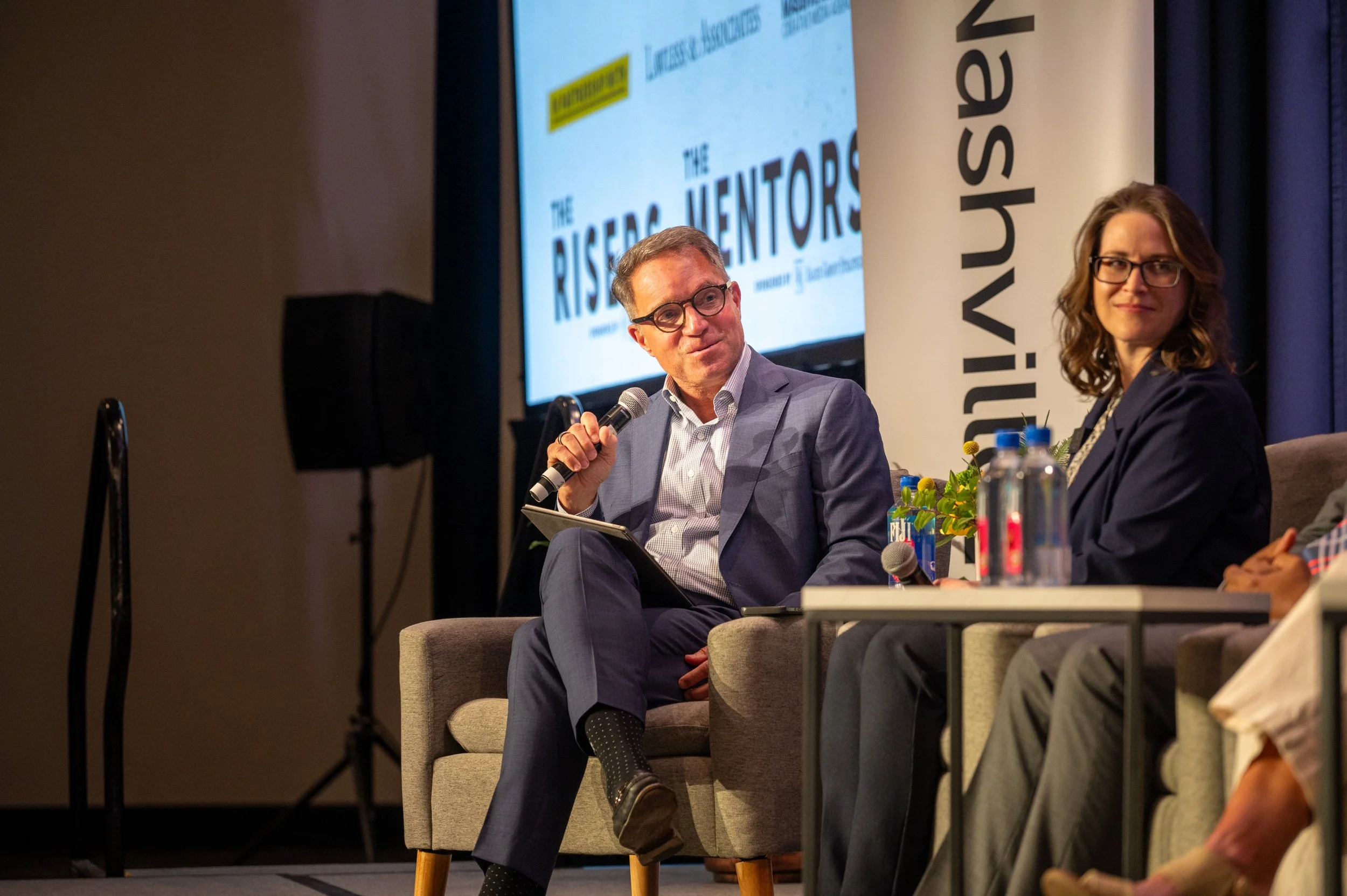 Two people sitting on a stage with a panel discussion setup. The man on the left is holding a microphone, wearing glasses and a light blue suit. The woman on the right is wearing glasses and a dark blazer. Behind them is a large screen displaying the