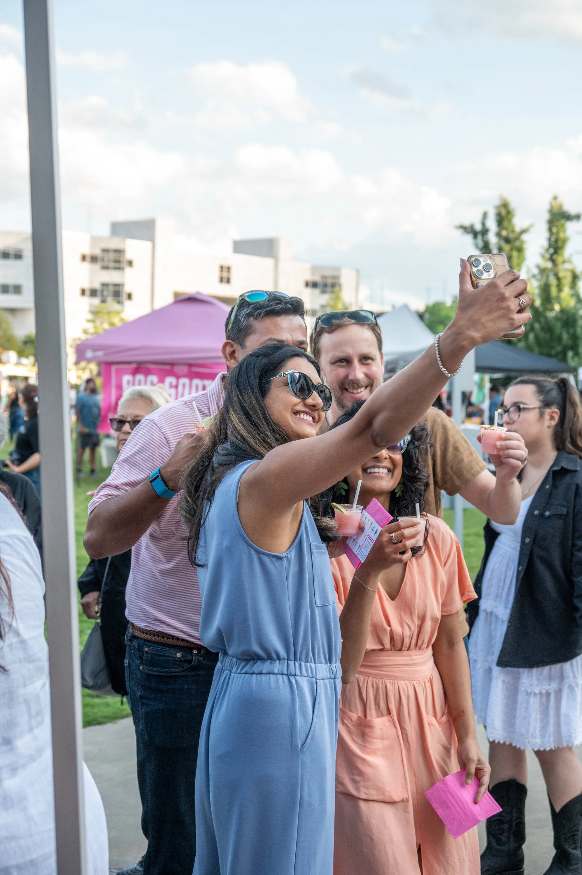 A group of people taking a selfie outdoors at a festival or fair, smiling and holding drinks.