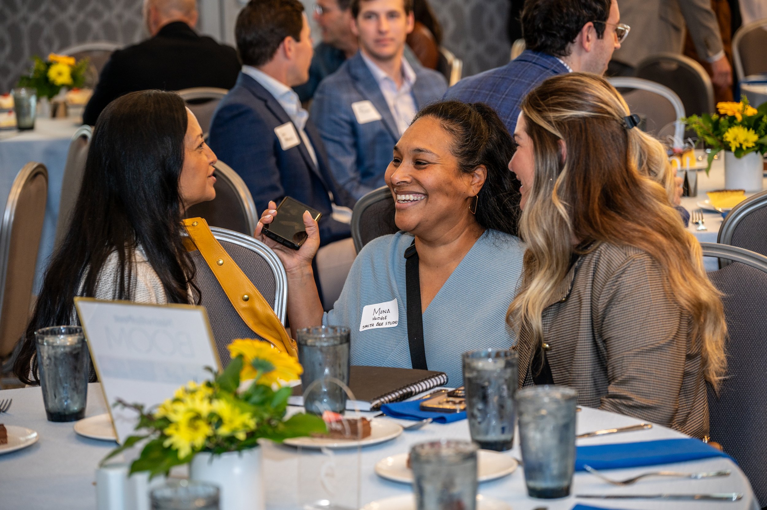 Women engaging in conversation at a professional event or conference, seated at a table with floral centerpiece, notebooks, and plates of dessert.