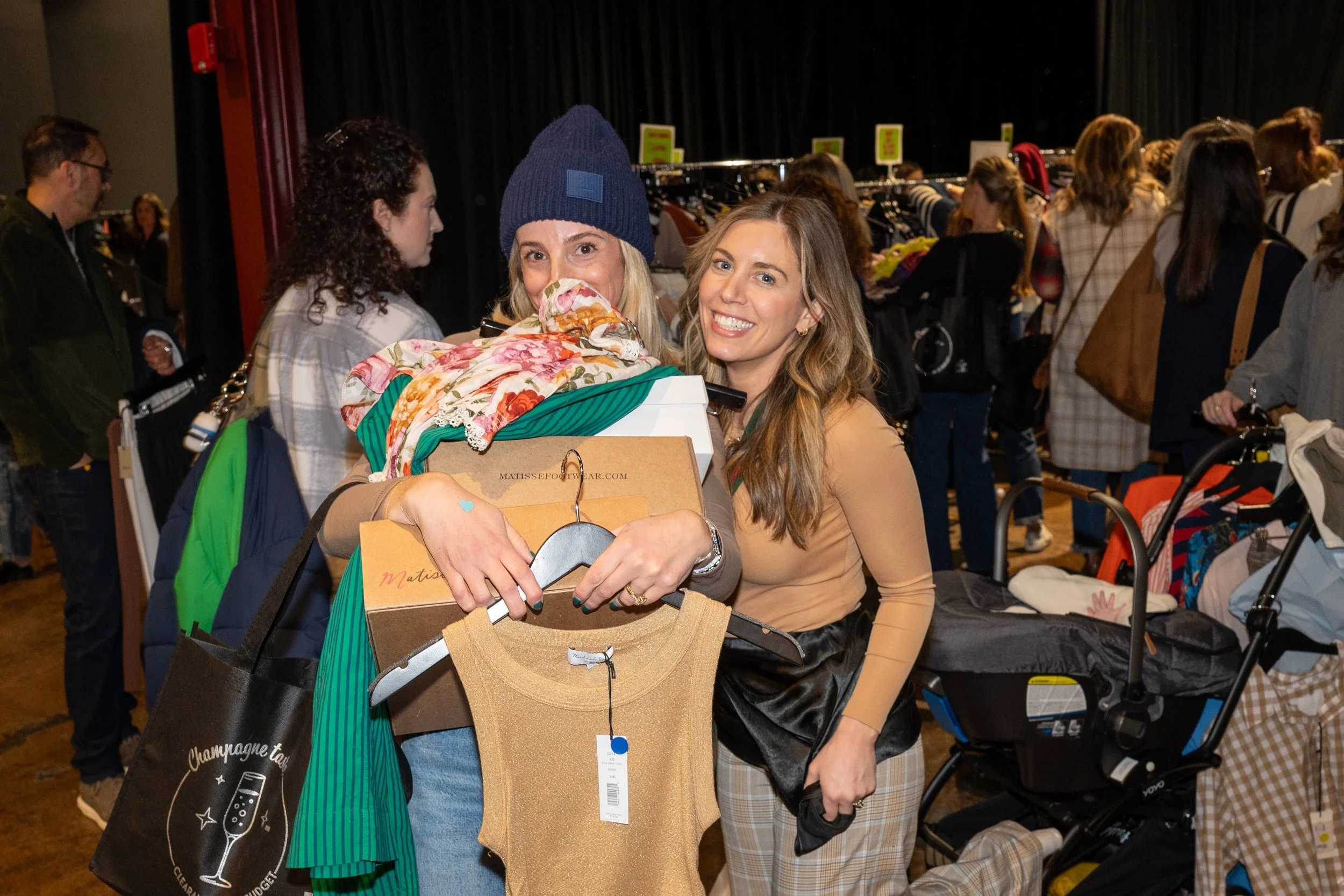 Two women smiling at a boutique warehouse sale, one holding clothes and a shopping bag, the other hugging her from the side.