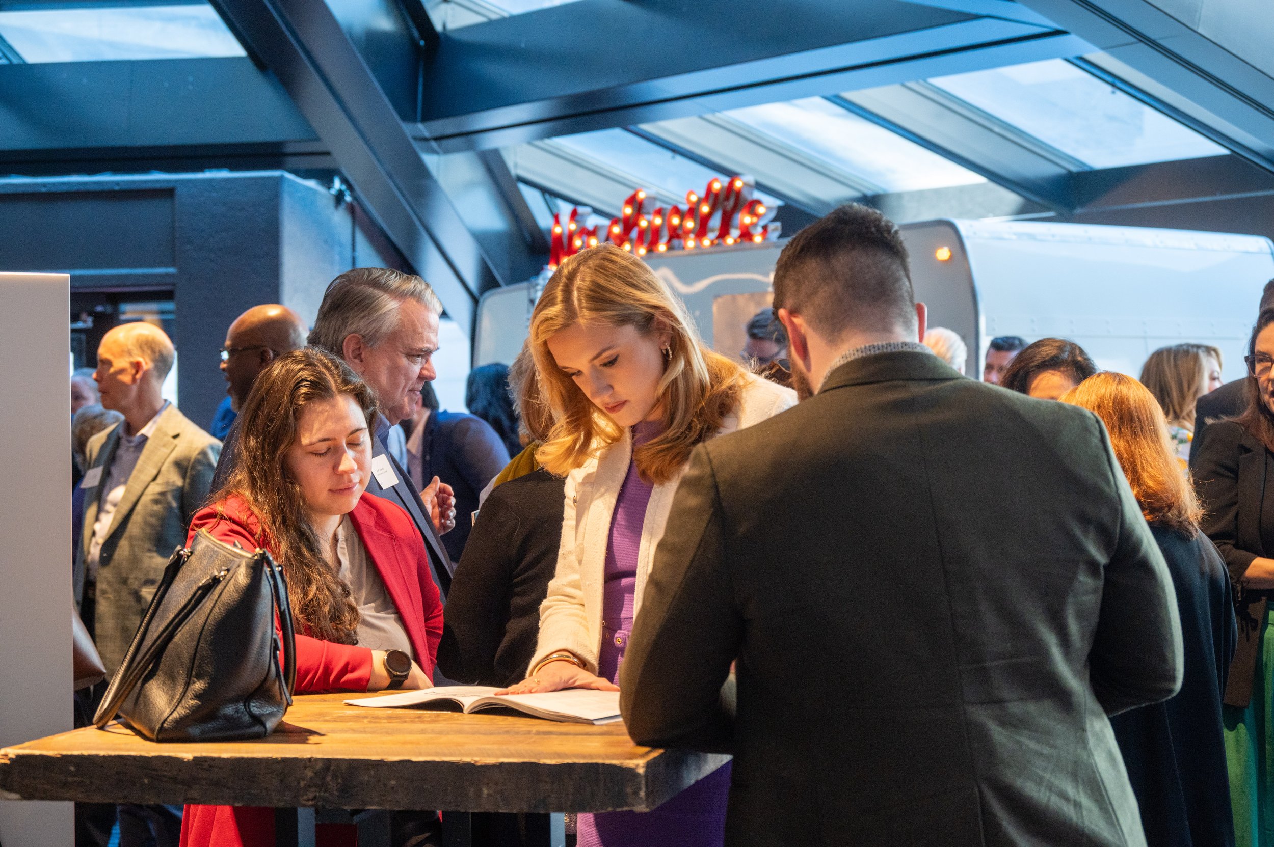 People gathered around a wooden table at a conference or event, some reading or writing, with a New Attic sign and modern architecture in the background.