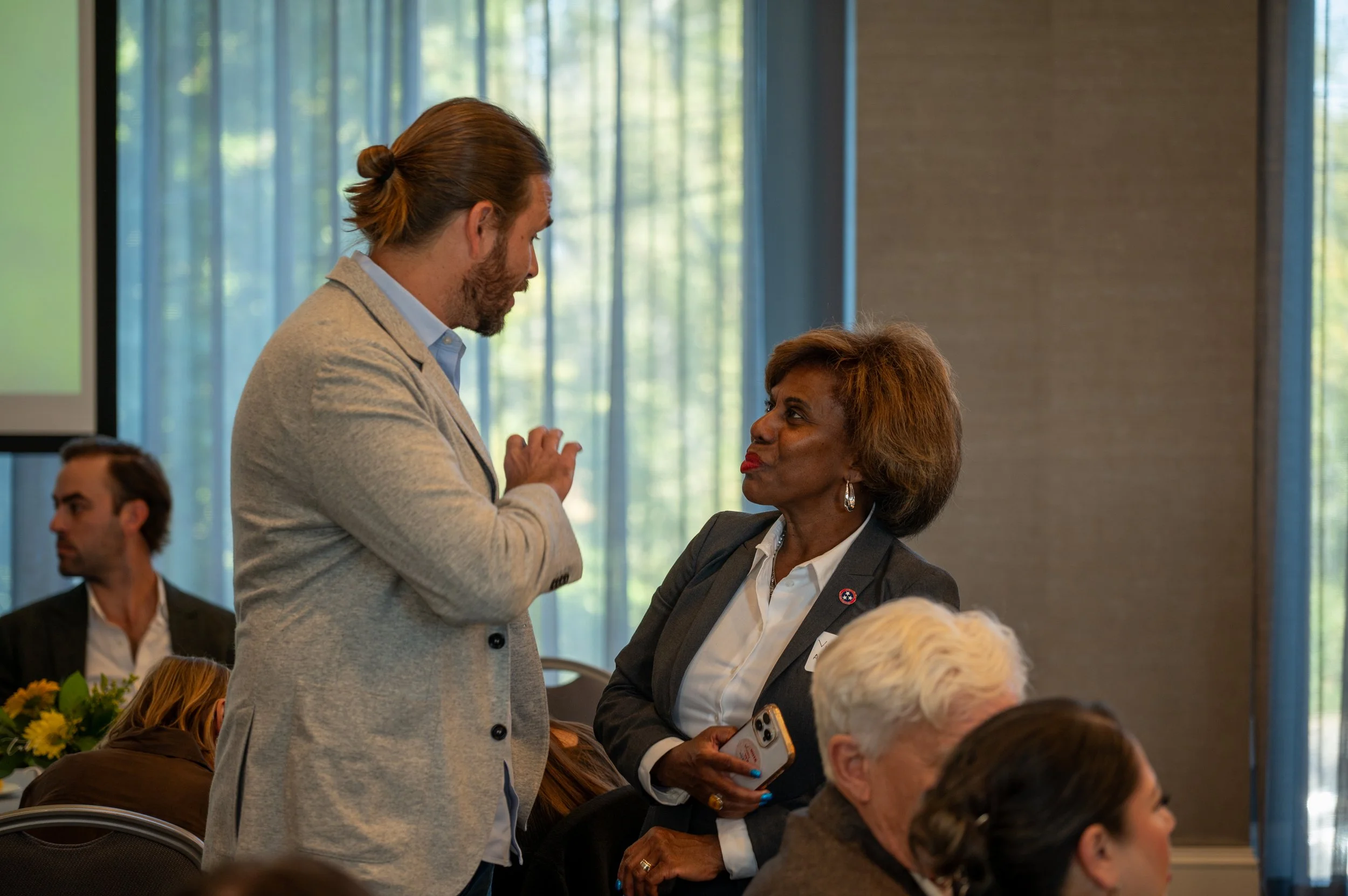 Two business professionals, a man and a woman, are engaged in a conversation during a conference or meeting in a well-lit room with large windows. The man has long hair tied back and is wearing a light gray blazer, while the woman has short hair and 