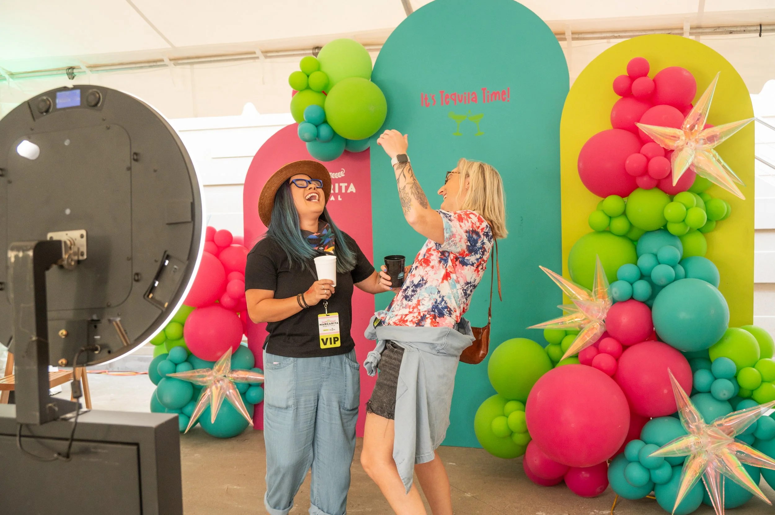 Two women laughing and dancing in front of colorful balloon decorations at a celebration event, with a sign saying "It's Tequila Time!" and a VIP badge.