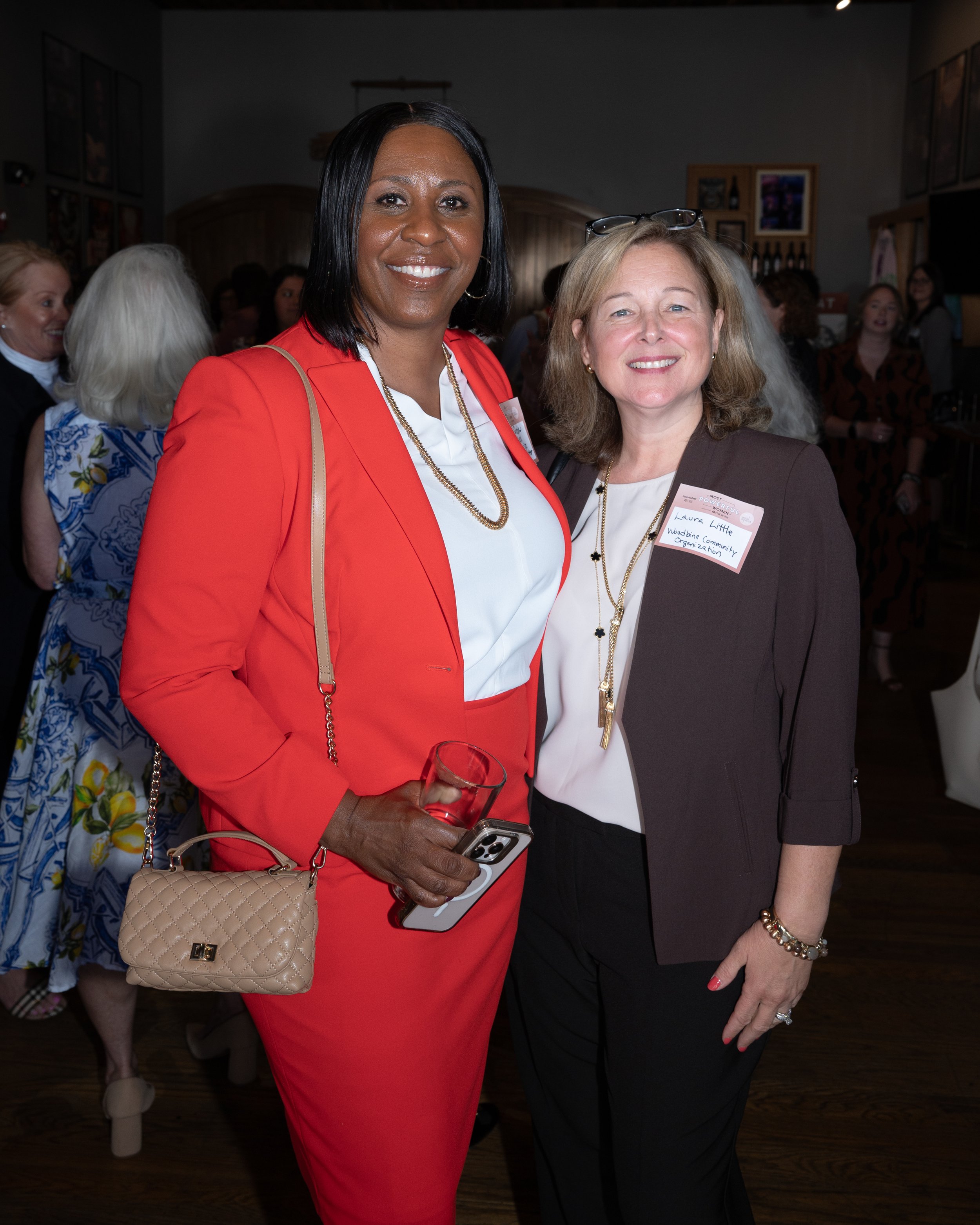 Two women standing close together at a social event, smiling at the camera. The woman on the left is wearing a red blazer and matching skirt with a white blouse, holding a glass, a phone, and a beige purse. The woman on the right is wearing a dark blazer and black pants