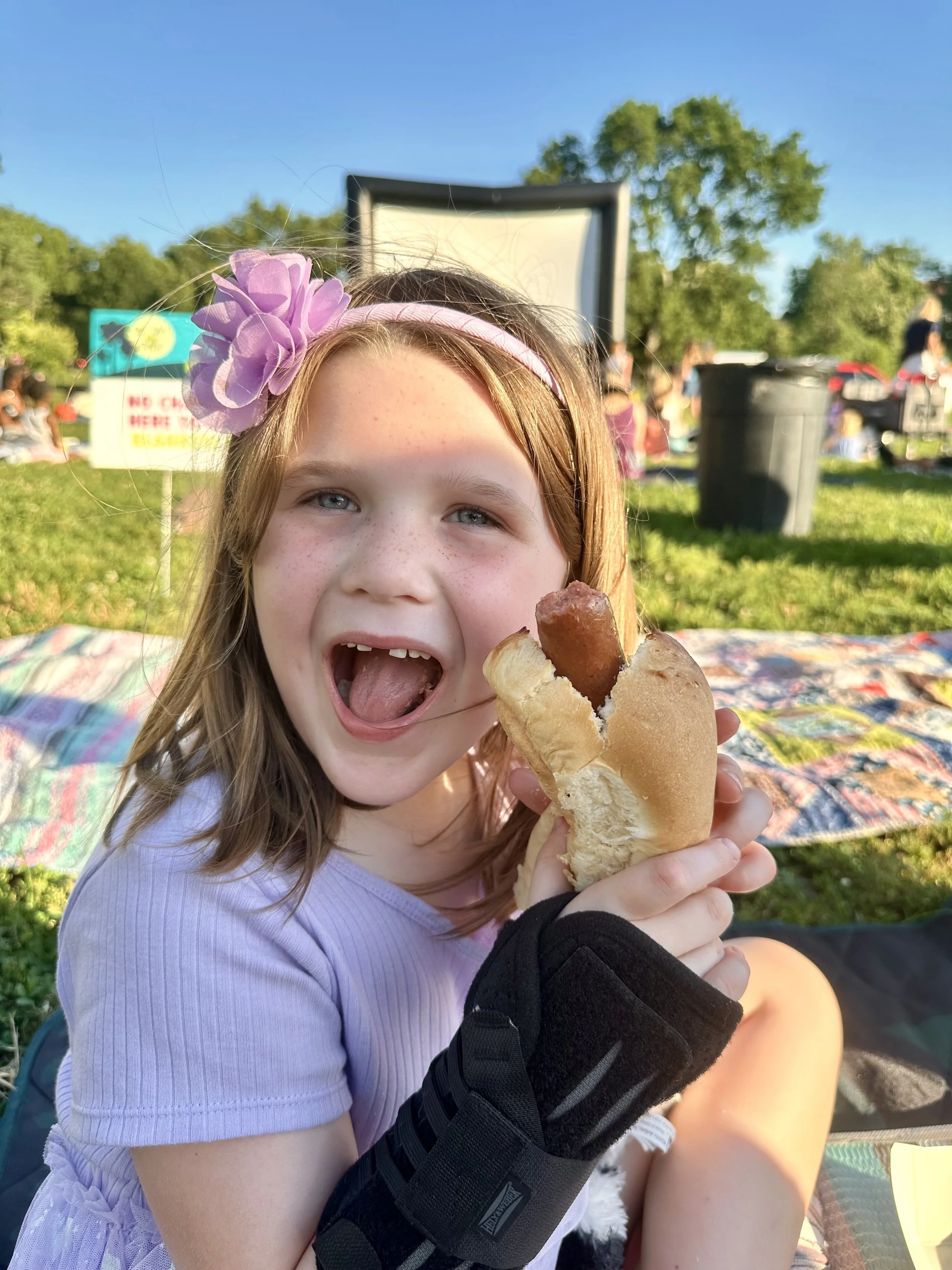 A young girl with red hair, wearing a purple shirt and a pink headband with a purple flower, is holding a hot dog with a sausage in a bun. She is smiling and has her mouth open, enjoying her food outdoors on a sunny day with green trees and people in the background.