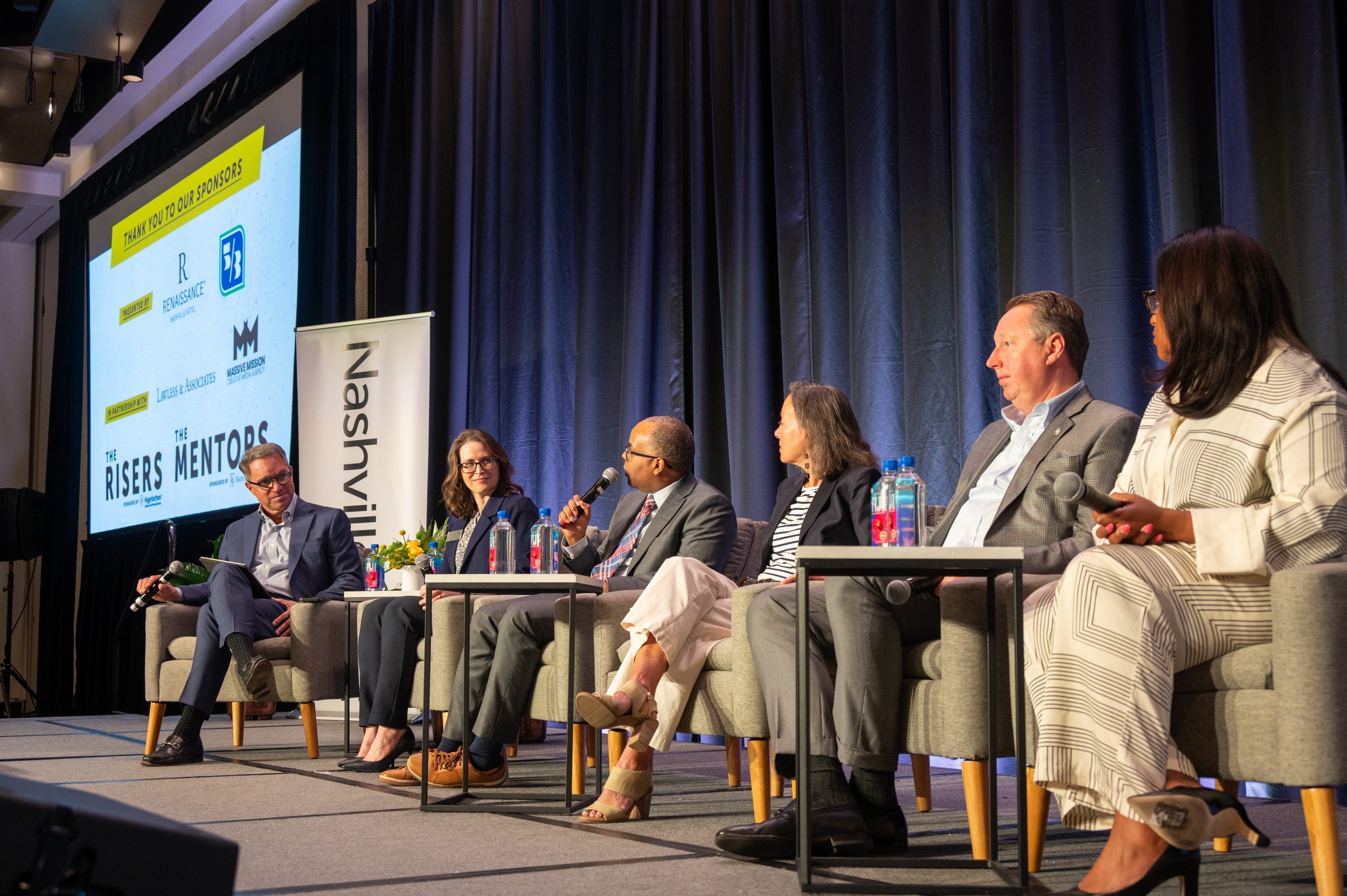 Six people sitting on a stage in chairs, participating in a panel discussion at a conference. Behind them is a large screen displaying sponsor logos and the event name 'The Risers Mentors'.