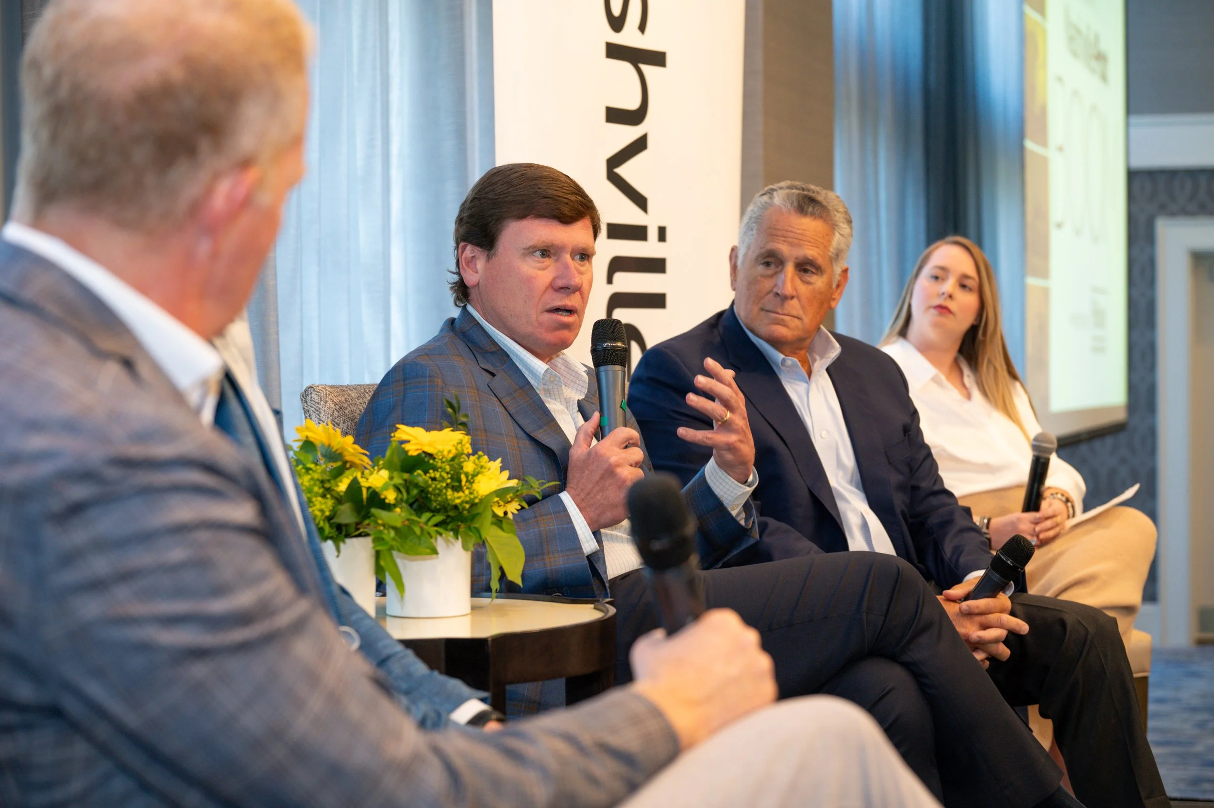 A panel of five people sitting on stage during a discussion or conference. The second man from the left is speaking into a microphone, and there are flowers on a table in front of him. There is a large banner with partially visible text behind them.