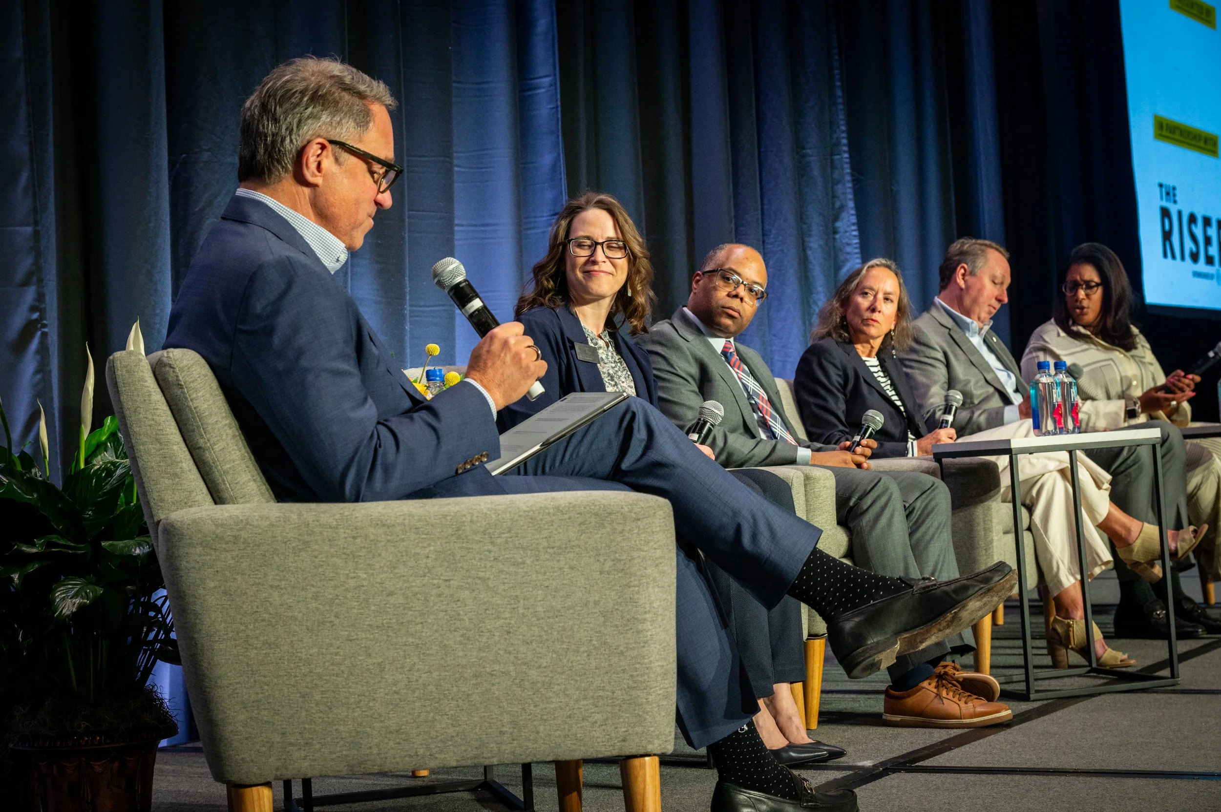 Six people sitting on stage at a panel discussion, one man on the far left holding a microphone and looking at a tablet, with five women and men next to him, some holding microphones and some using their phones.