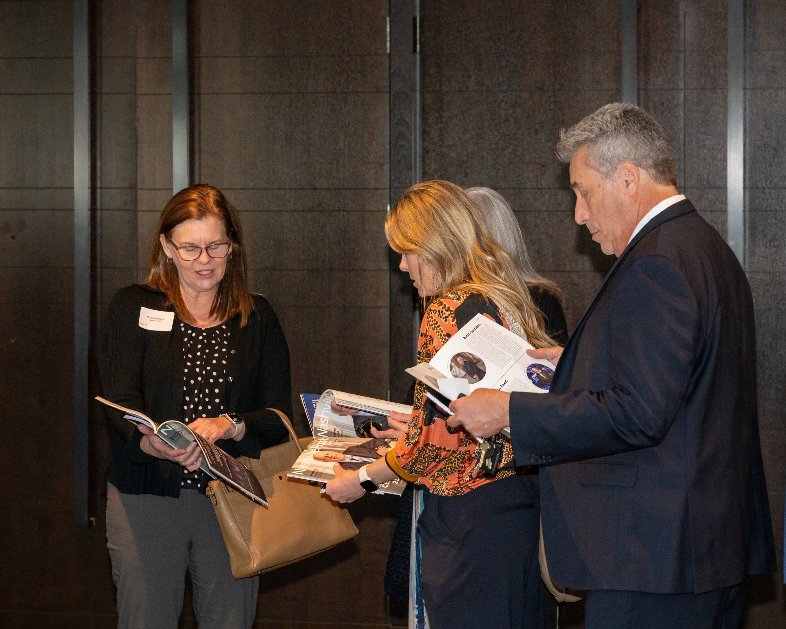 A group of four business professionals, three women and one man, standing close together and looking at magazines or brochures in an indoor setting with a dark wall background.