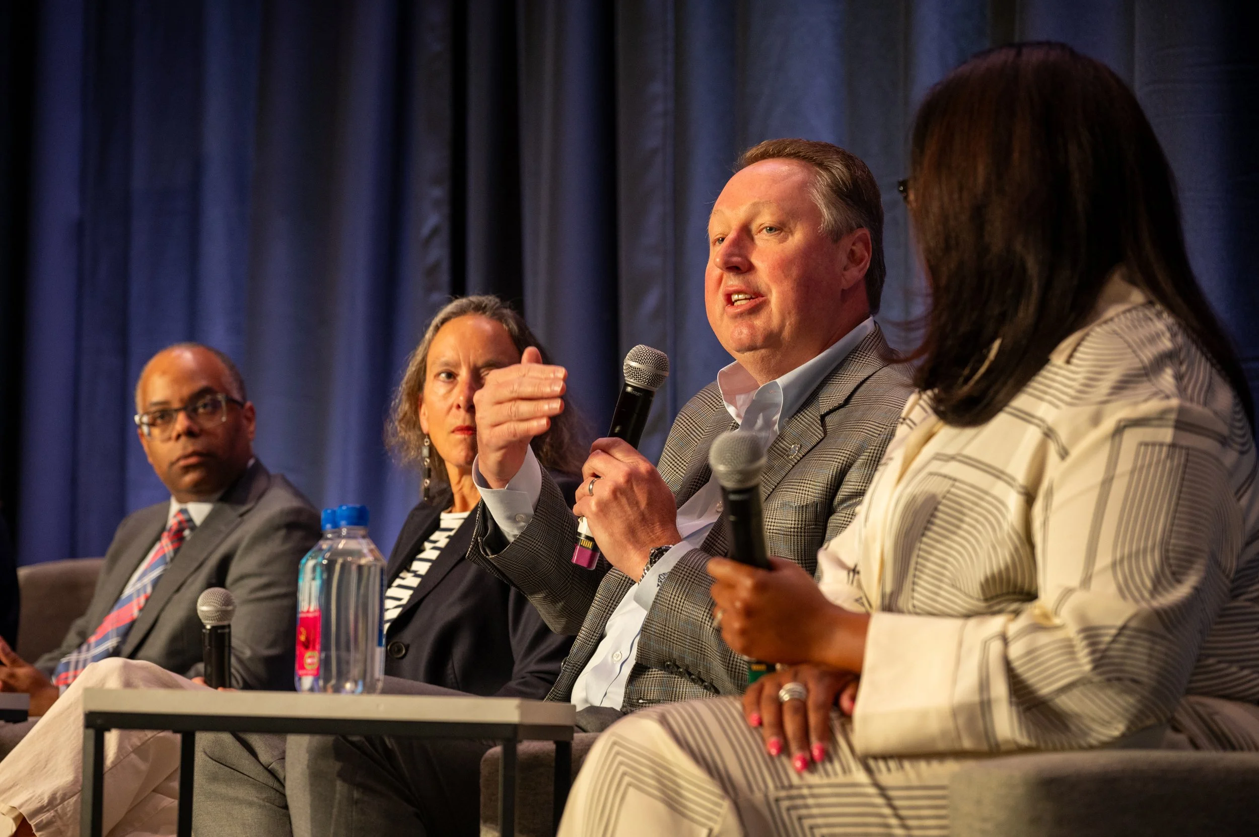 Panel of four diverse individuals speaking into microphones at a conference or seminar, seated on stage with a dark curtain backdrop.