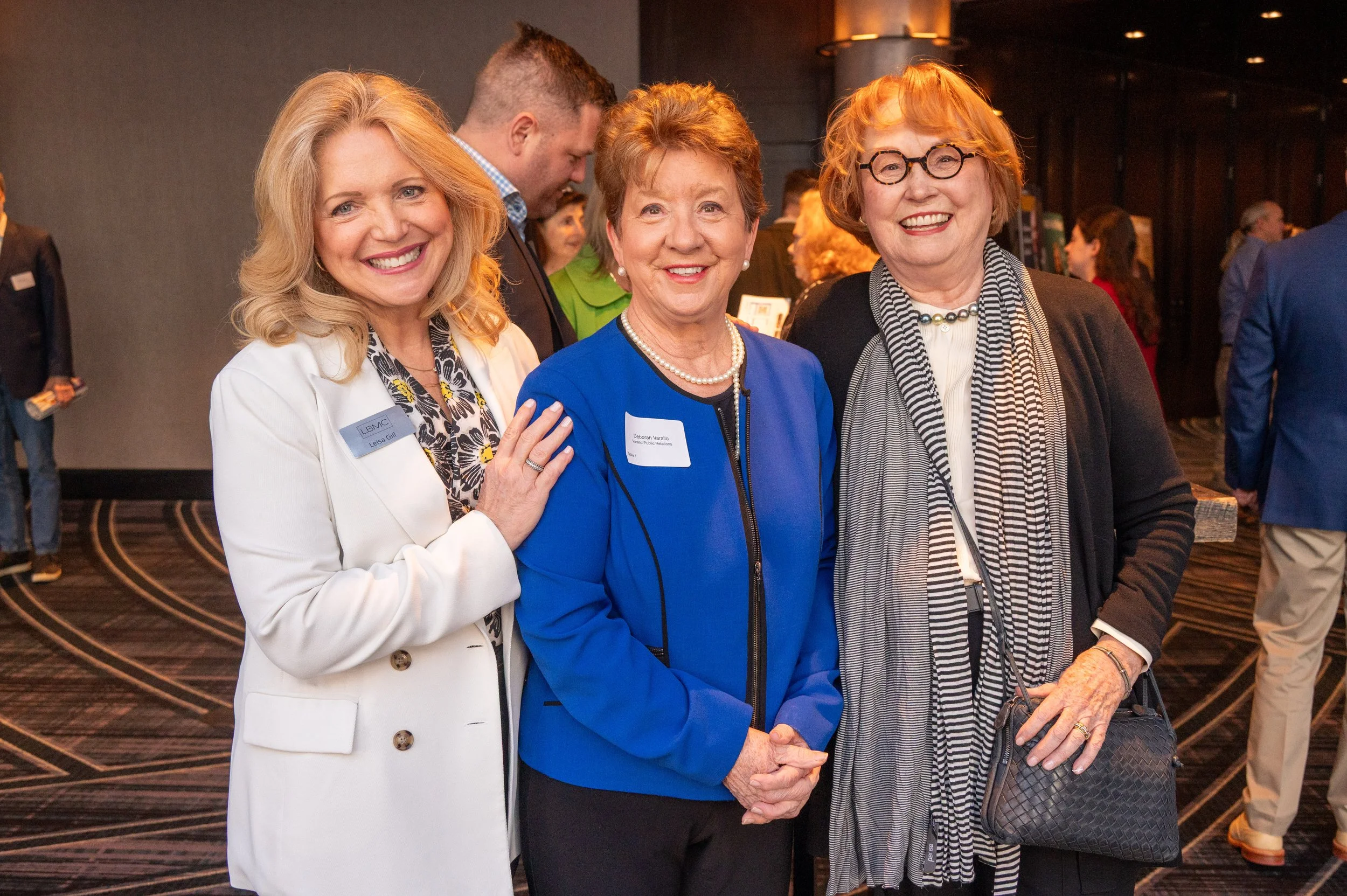 Three women standing together at an indoor event, smiling at the camera. The woman on the left has blonde hair, is wearing a white blazer, and a name tag. The woman in the middle has short, curly reddish hair, and is dressed in a blue blazer with a p