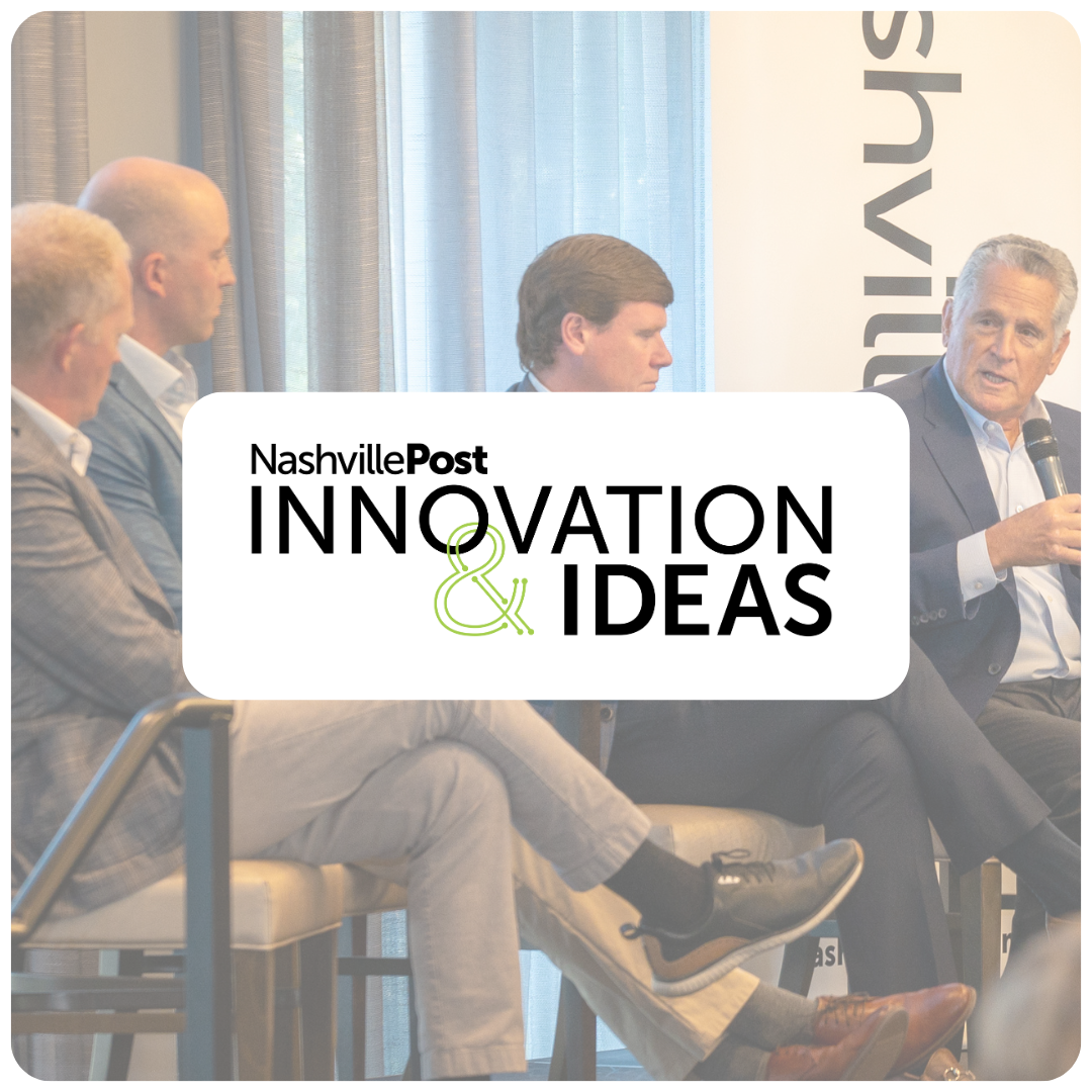 Panel discussion at Nashville Post Innovation & Ideas event with four men seated, one speaking into a microphone, in an indoor setting with curtains and a screen in the background.