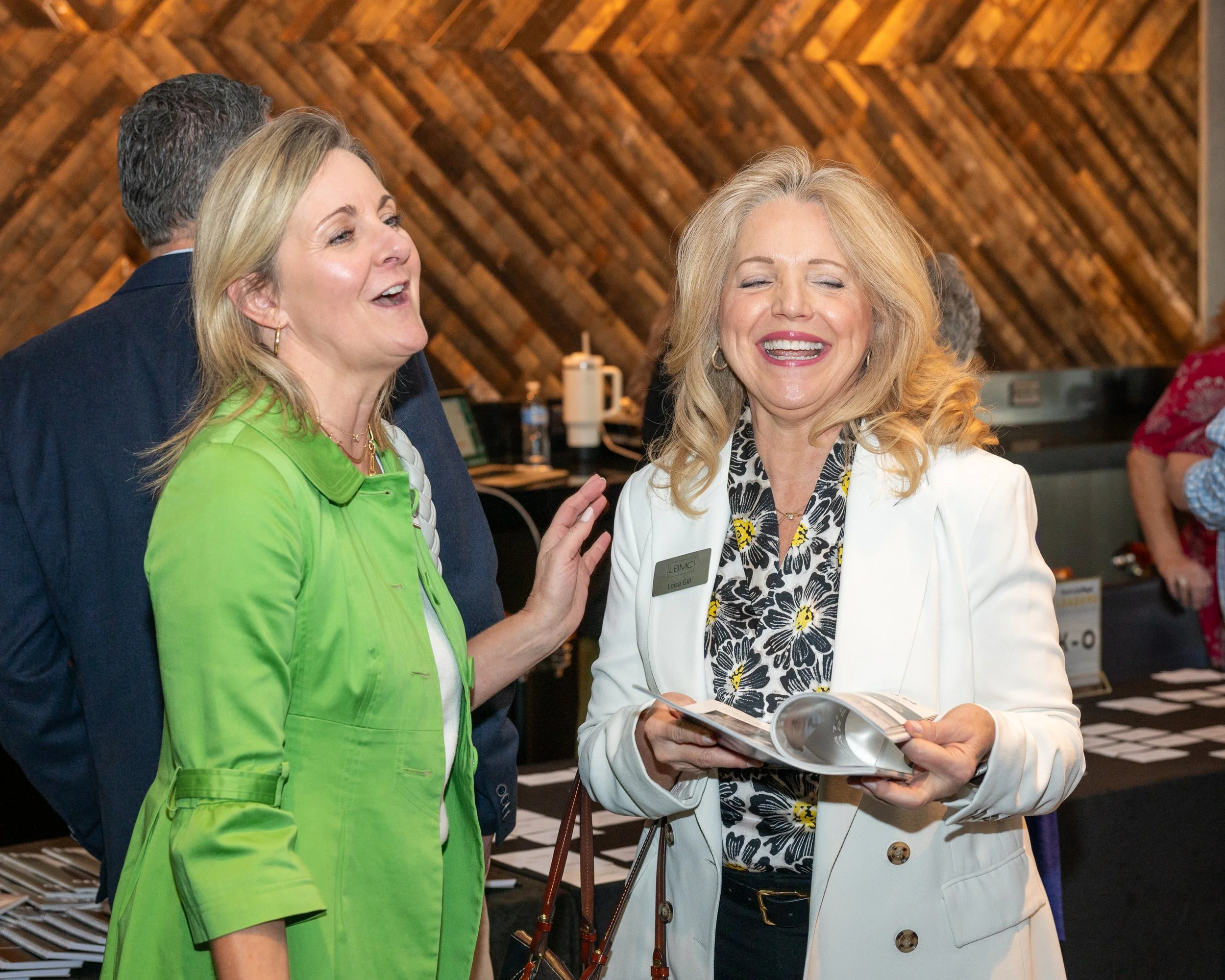 Two women laughing and engaging in conversation at an indoor event, with one wearing a white blazer and the other a bright green jacket, in front of a wooden wall background.