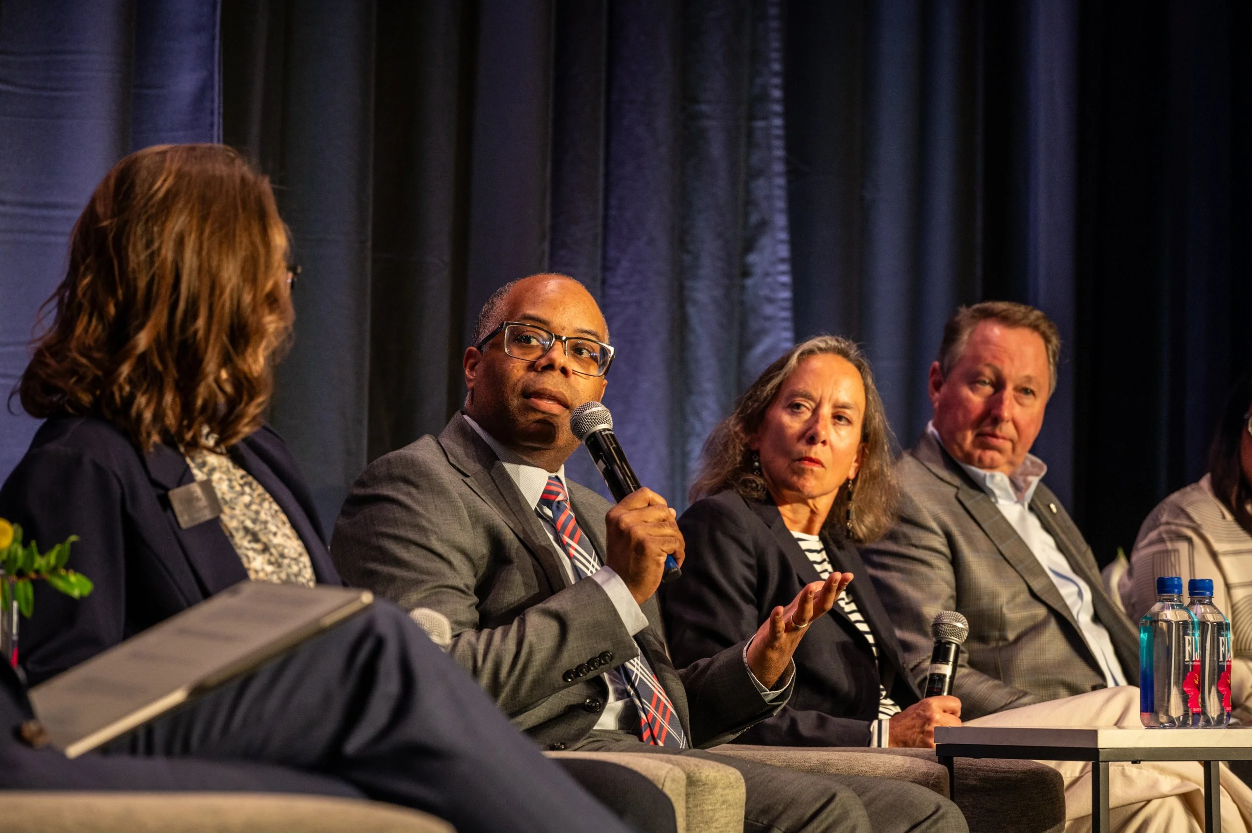 A group of people sitting on a panel at a conference, engaged in discussion, with one man speaking into a microphone, against a dark curtain background.