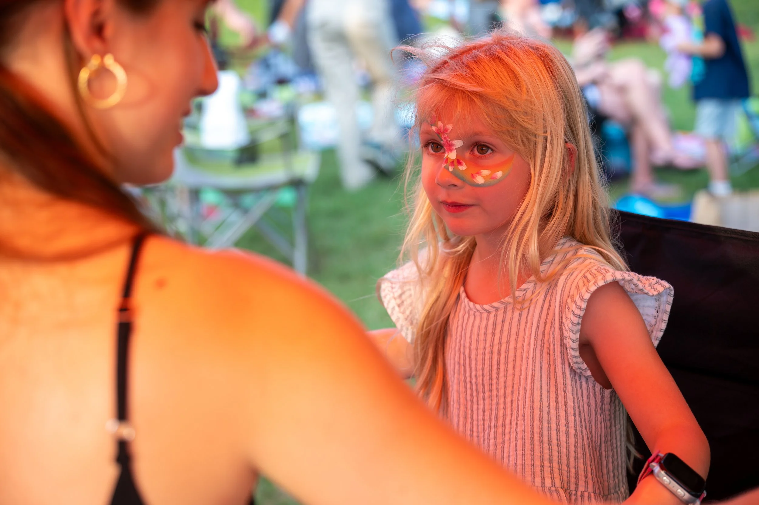 A young girl with face paint of a butterfly and flowers looking at an adult woman at an outdoor event.