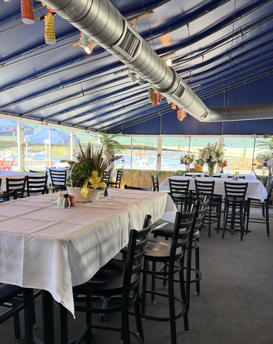 Indoor outdoor dining area with tables covered in white tablecloths, decorated with centerpieces of flowers and greenery, set up for an event or gathering, with large windows, blue ceiling, and colorful hanging decorations.