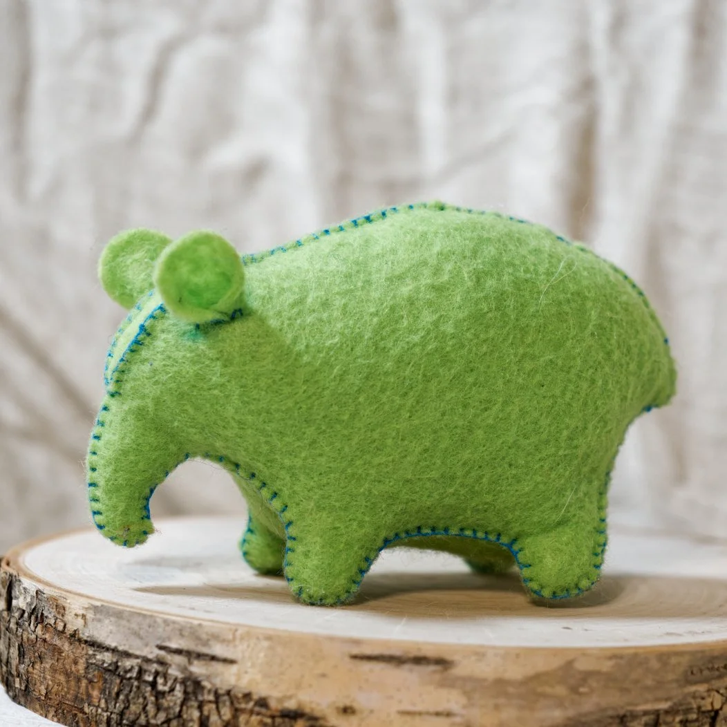 Green felt plush toy in the shape of a tapir, with small round ears, placed on a wooden surface with a textured white background.