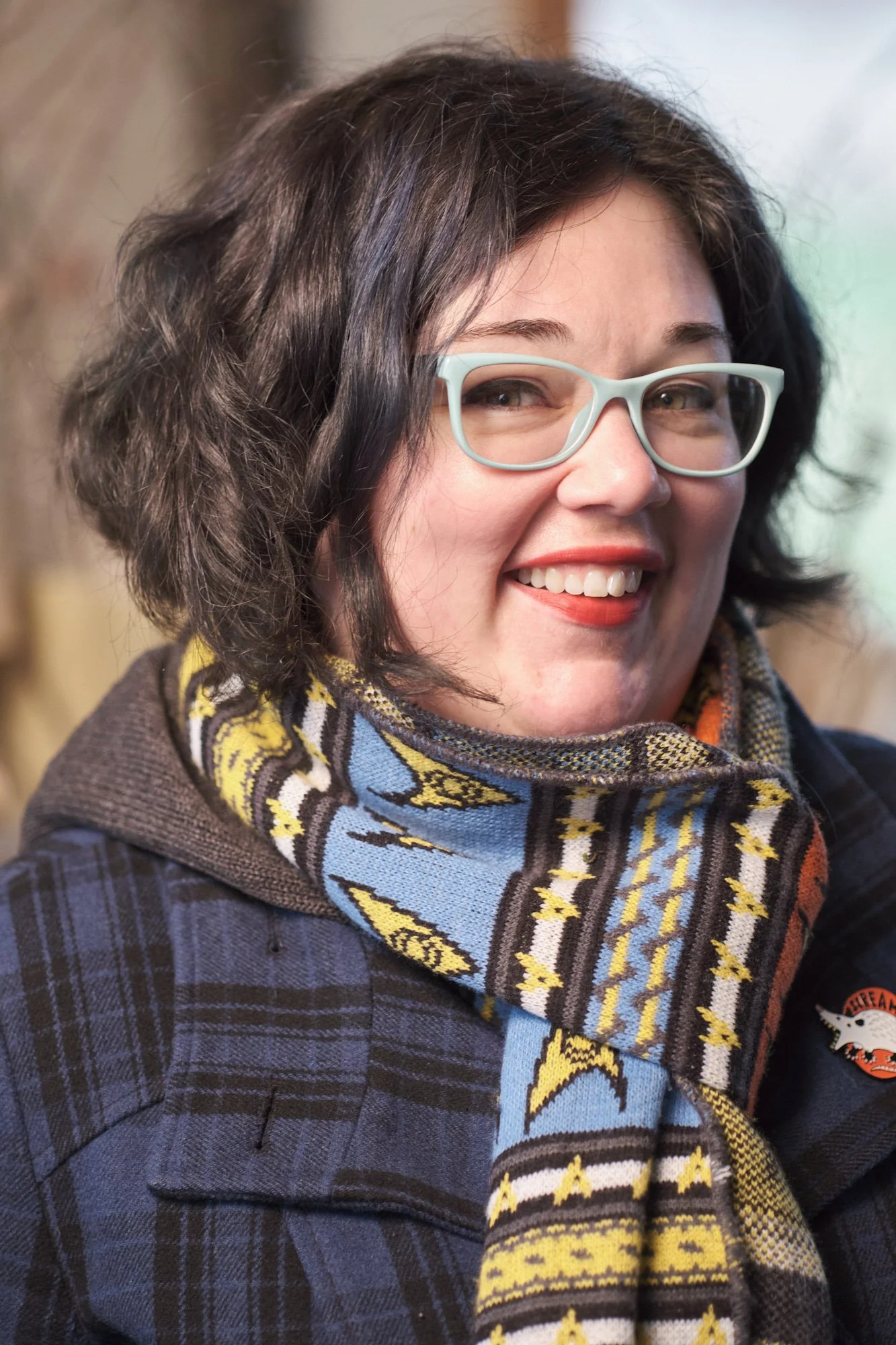 Angela, a woman with curly dark hair, light-colored glasses, and a colorful scarf, smiling at the camera.