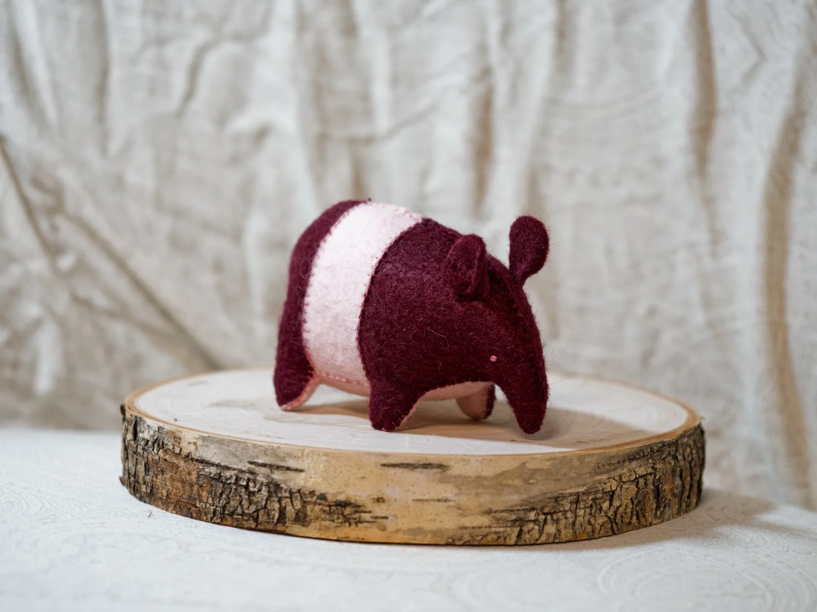 A felt toy in the shape of a tapir, with a burgundy and pink color, resting on a wooden slab.