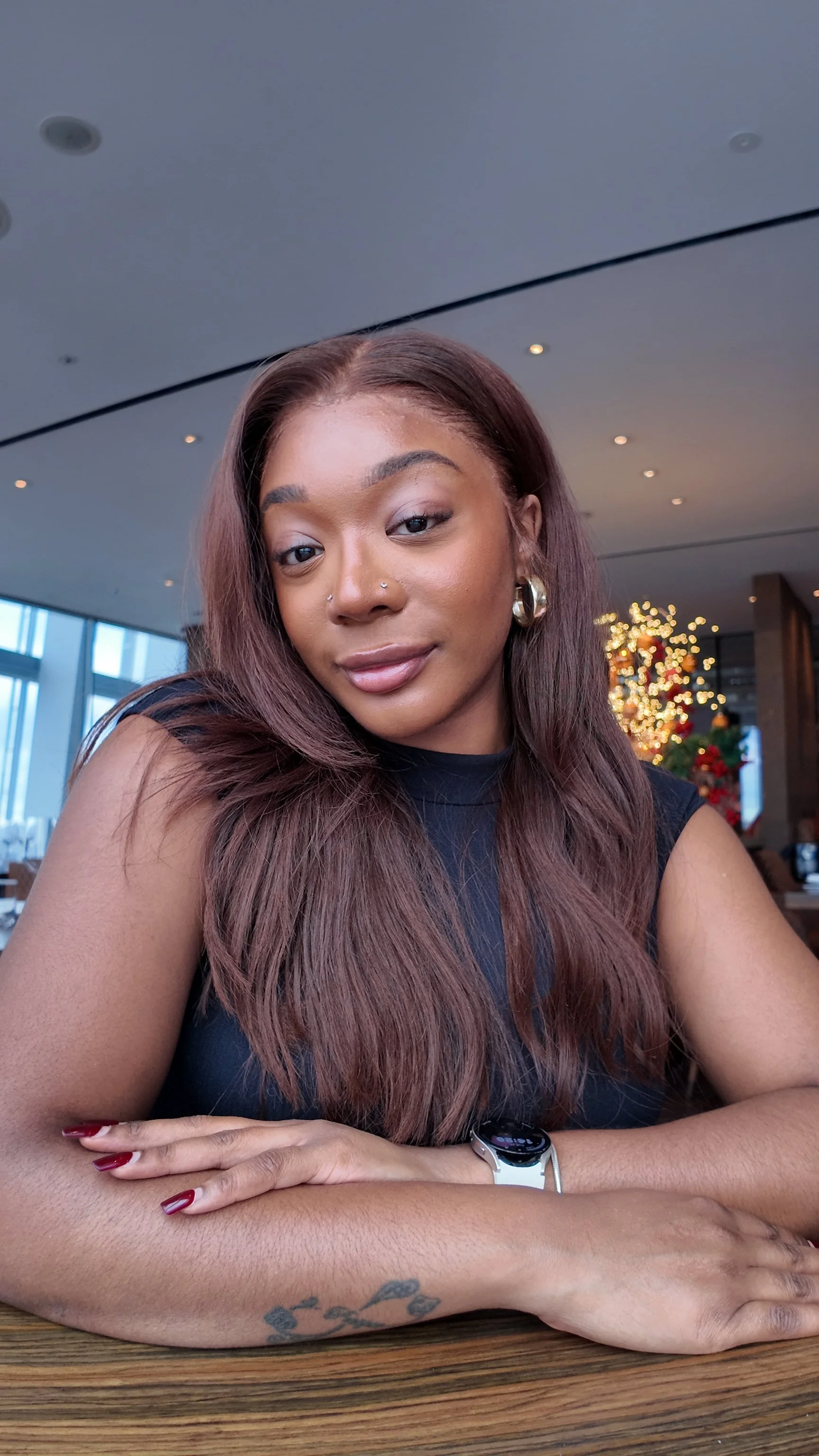 A woman with long wavy hair, wearing a black sleeveless top, sitting at a table in a decorated indoor space with Christmas lights and a Christmas tree in the background.