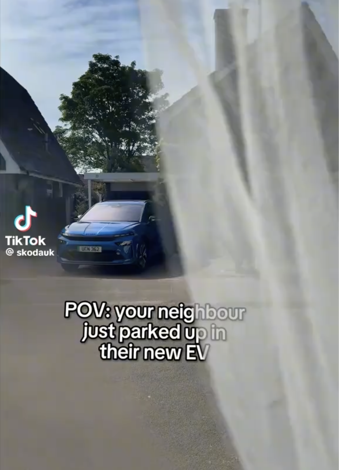 A blue electric vehicle (EV) parked in a driveway with a tree and houses in the background, viewed through a semi-transparent curtain or fabric.