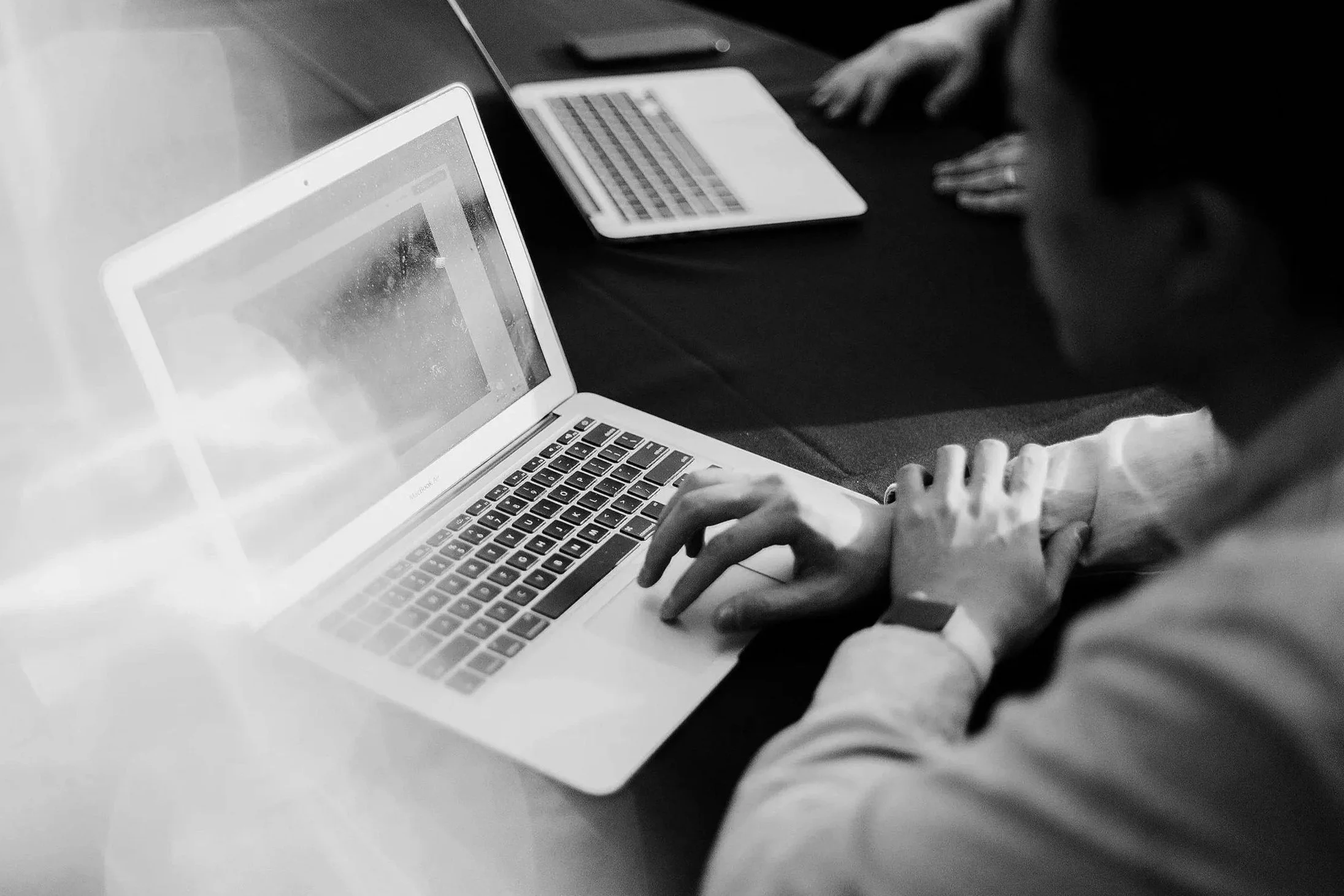 Person working on a laptop at a table with another open laptop and a tablet in the background.