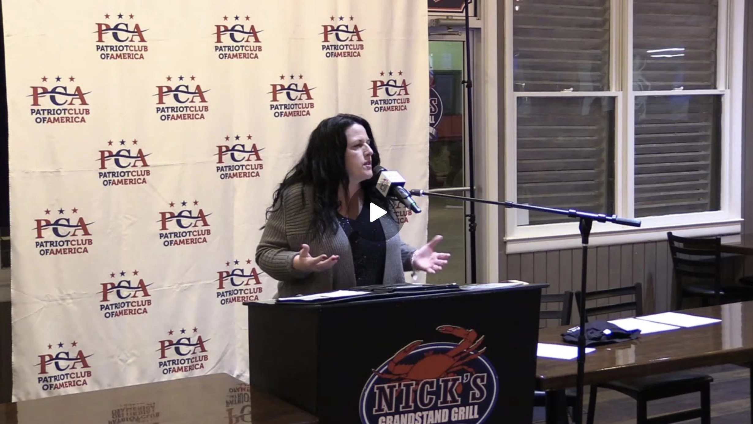 A woman with long dark hair speaking at a podium with a microphone, in front of a backdrop with the logo of Patriot Club of America. The podium has a sign for Nick's Grandstand Grill. There are chairs and tables nearby, with papers and a phone on the table. Large windows are behind her.