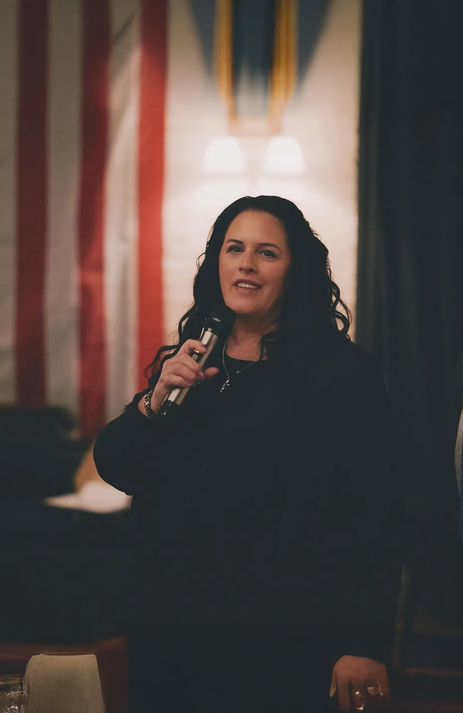 A woman with long dark hair holding a microphone is speaking at an indoor event, with soft lighting and decorative curtains in the background.