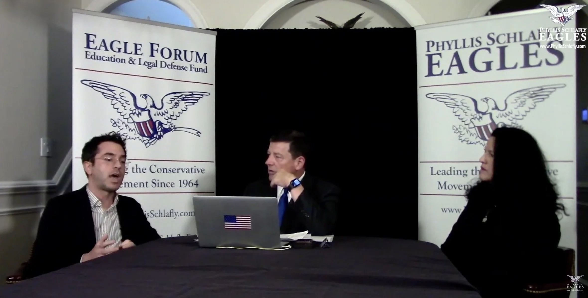 Three people seated at a table during a discussion or interview, with banners in the background displaying the logos and name of the Phyllis Schlafly Eagles and the Eagle Forum, organization titles, and slogans.