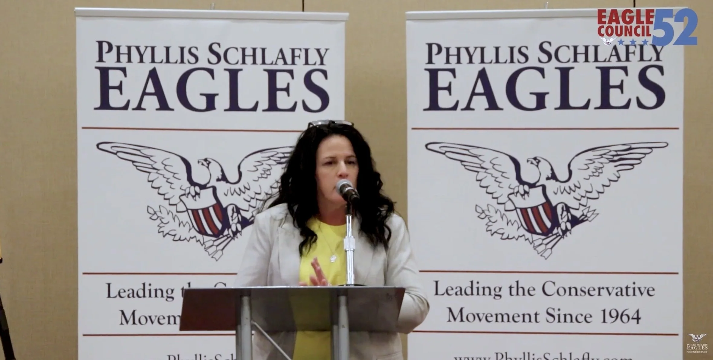 A woman with dark curly hair speaking into a microphone at a podium during a political event with two banners behind her. The banners read 'Phyllis Schlafly Eagles' and 'Leading the Conservative Movement Since 1964'. The event appears to be affiliated with Eagle Council 52.