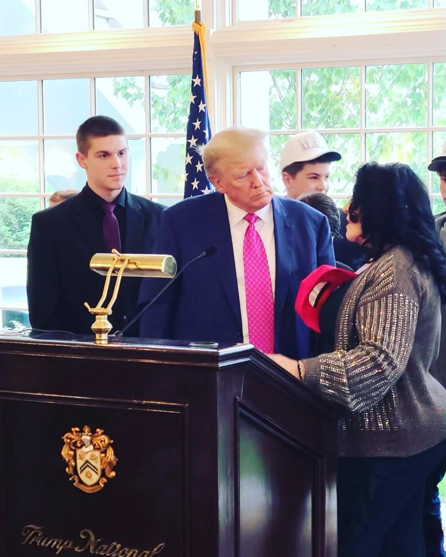 Donald Trump standing at a podium with a woman in front of him, surrounded by young people, in a room with large windows and an American flag in the background.