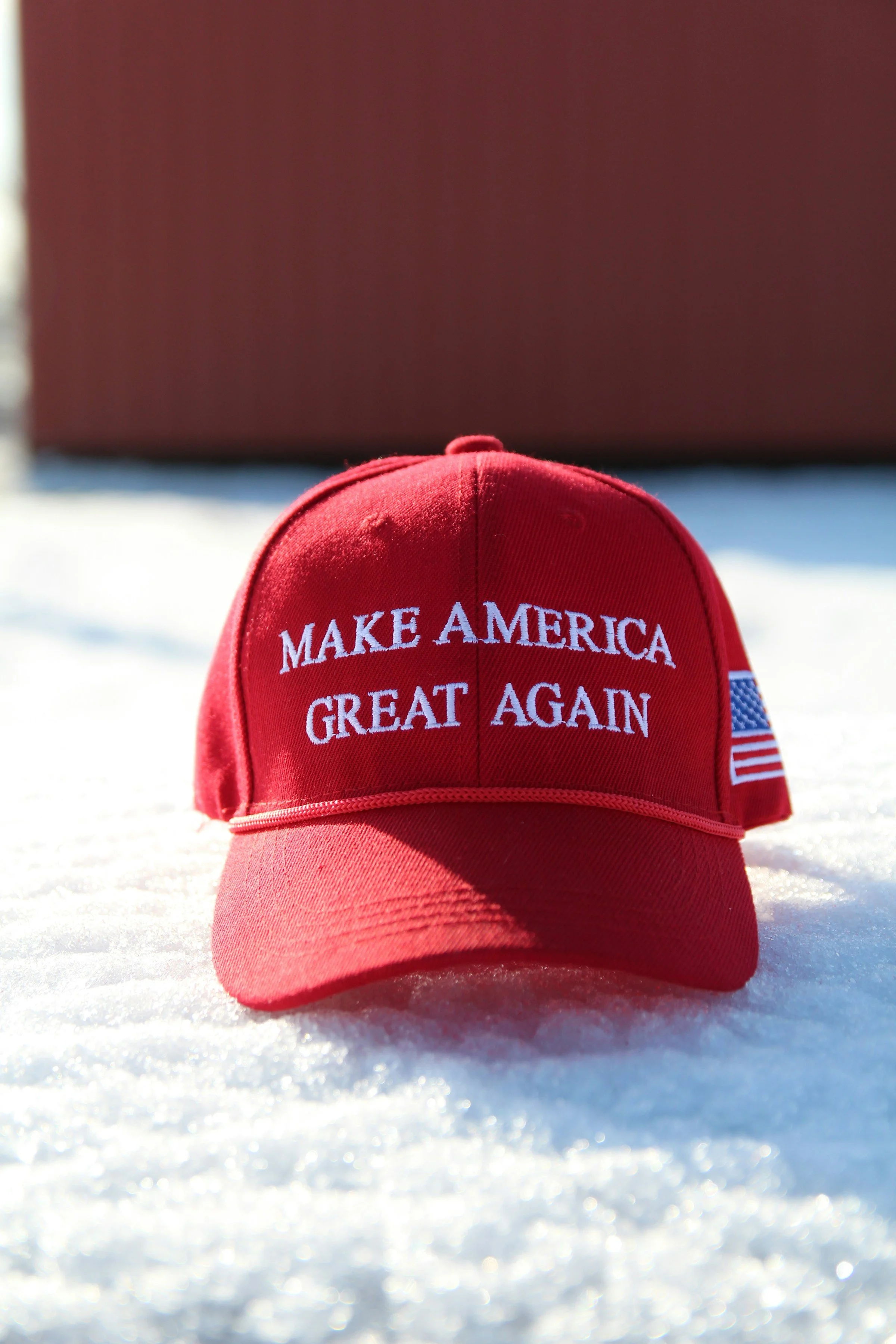 Red baseball cap with 'MAKE AMERICA GREAT AGAIN' embroidered in white, placed on snow with a building in the background.