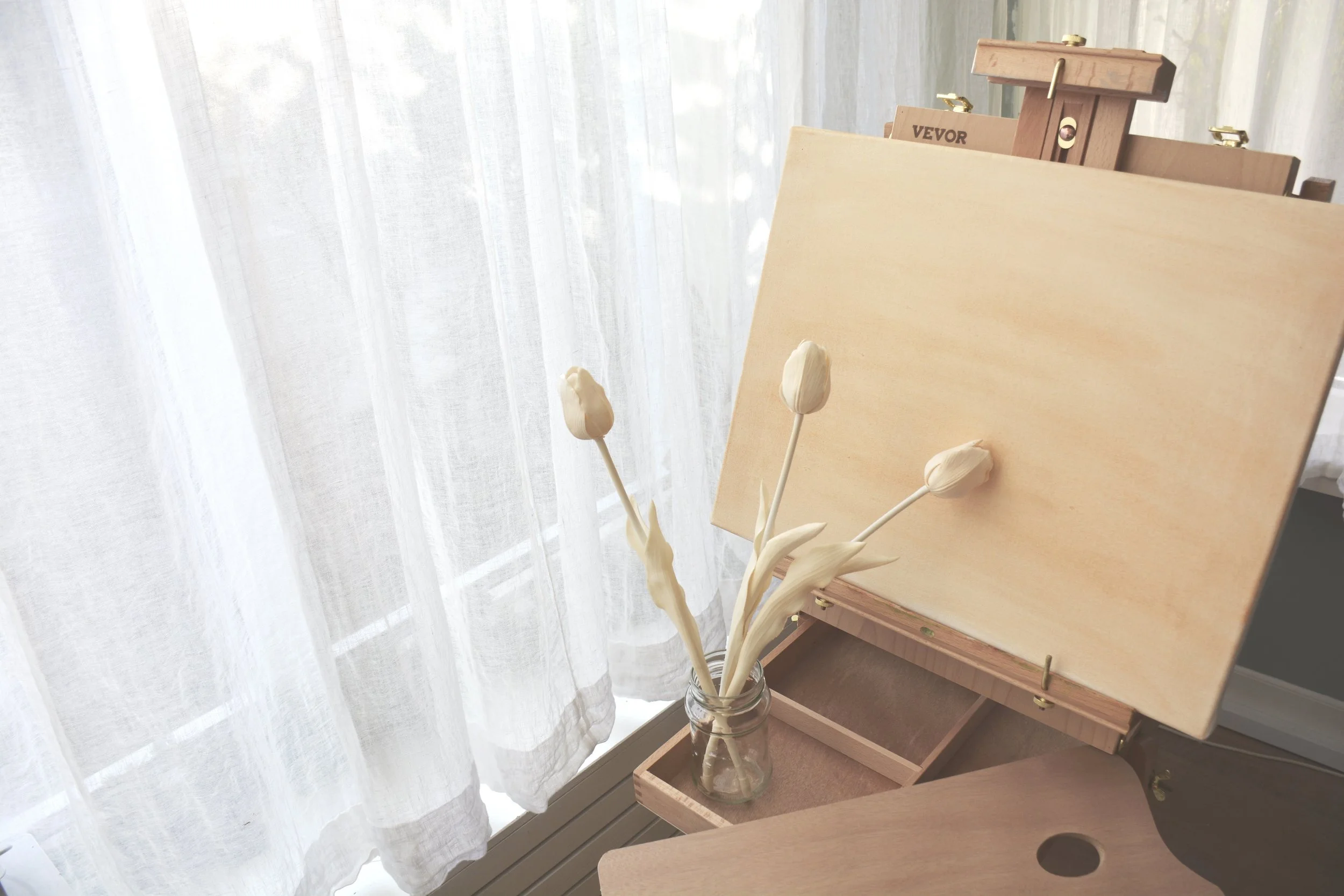 Empty wooden artist's easel, blank canvas, and a small jar with white tulips in front of sheer white curtains.