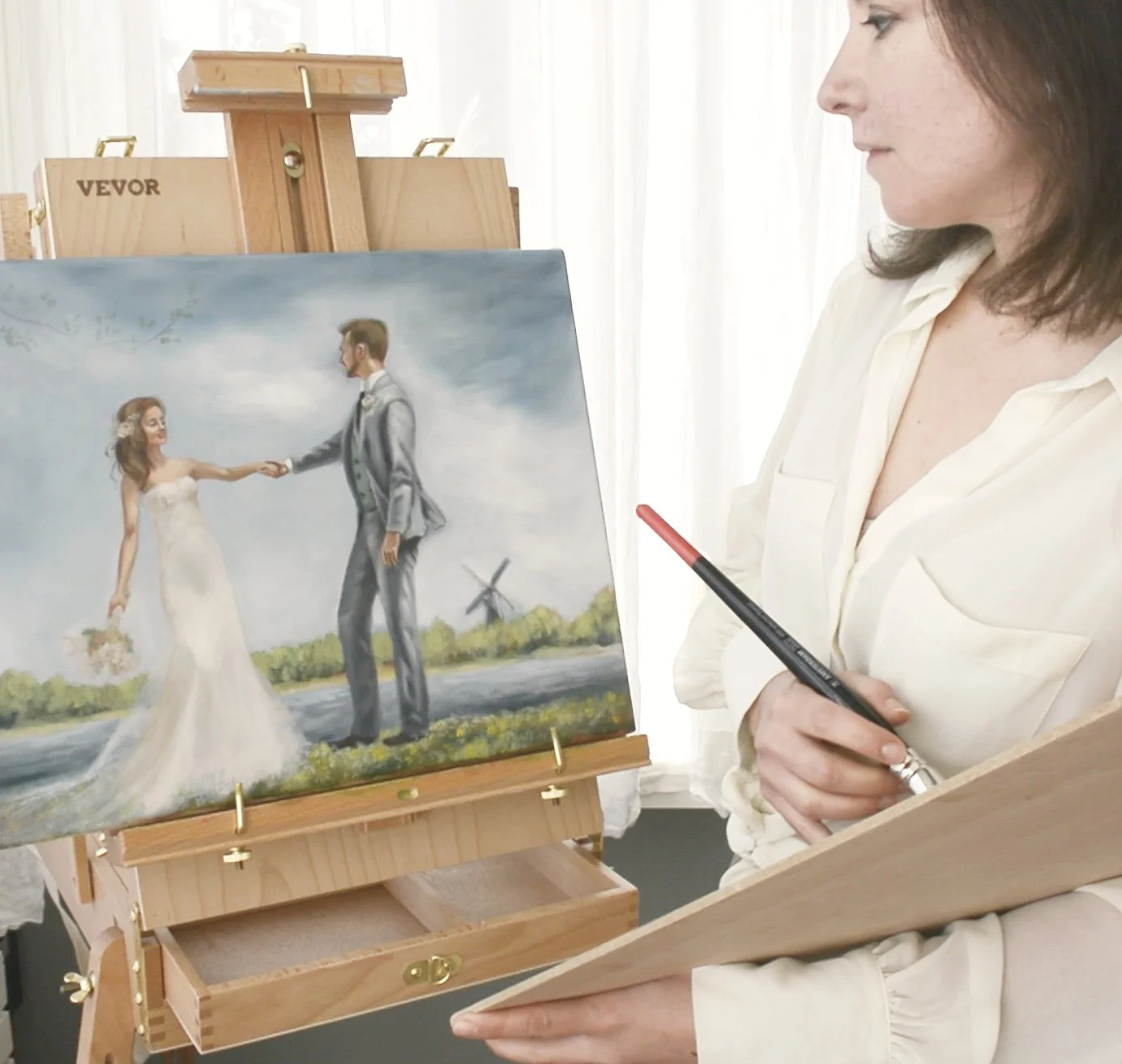 The image shows a woman holding a paintbrush and a palette, working on a wedding-themed painting of a bride and groom holding hands near a river with a windmill in the background.