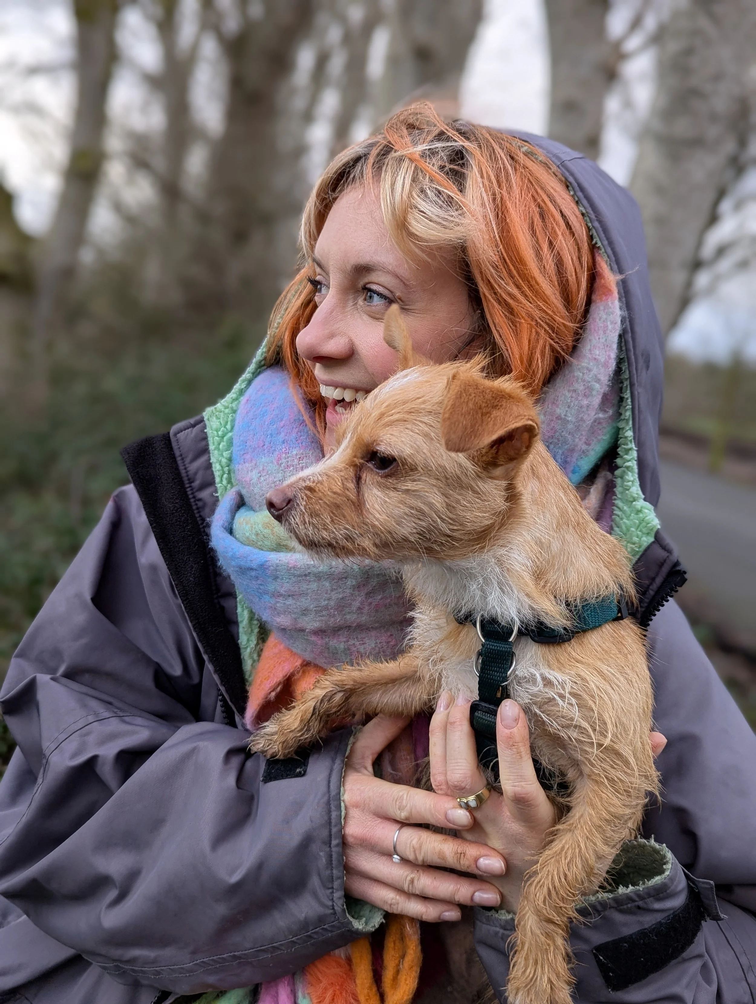A woman with red hair in a colorful scarf and a gray jacket is holding a small brown dog with a black collar outdoors, with trees in the background.