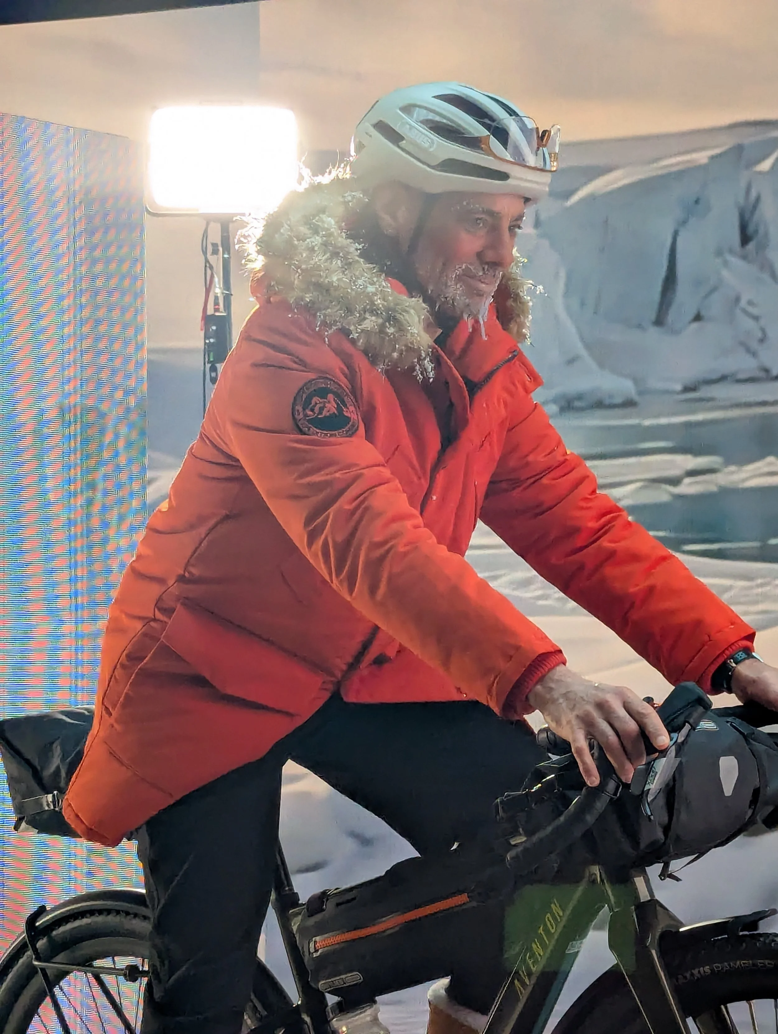 Man riding a bicycle indoors, wearing a white helmet, orange winter jacket with a fur-lined hood, and black pants, with a bright light behind him.