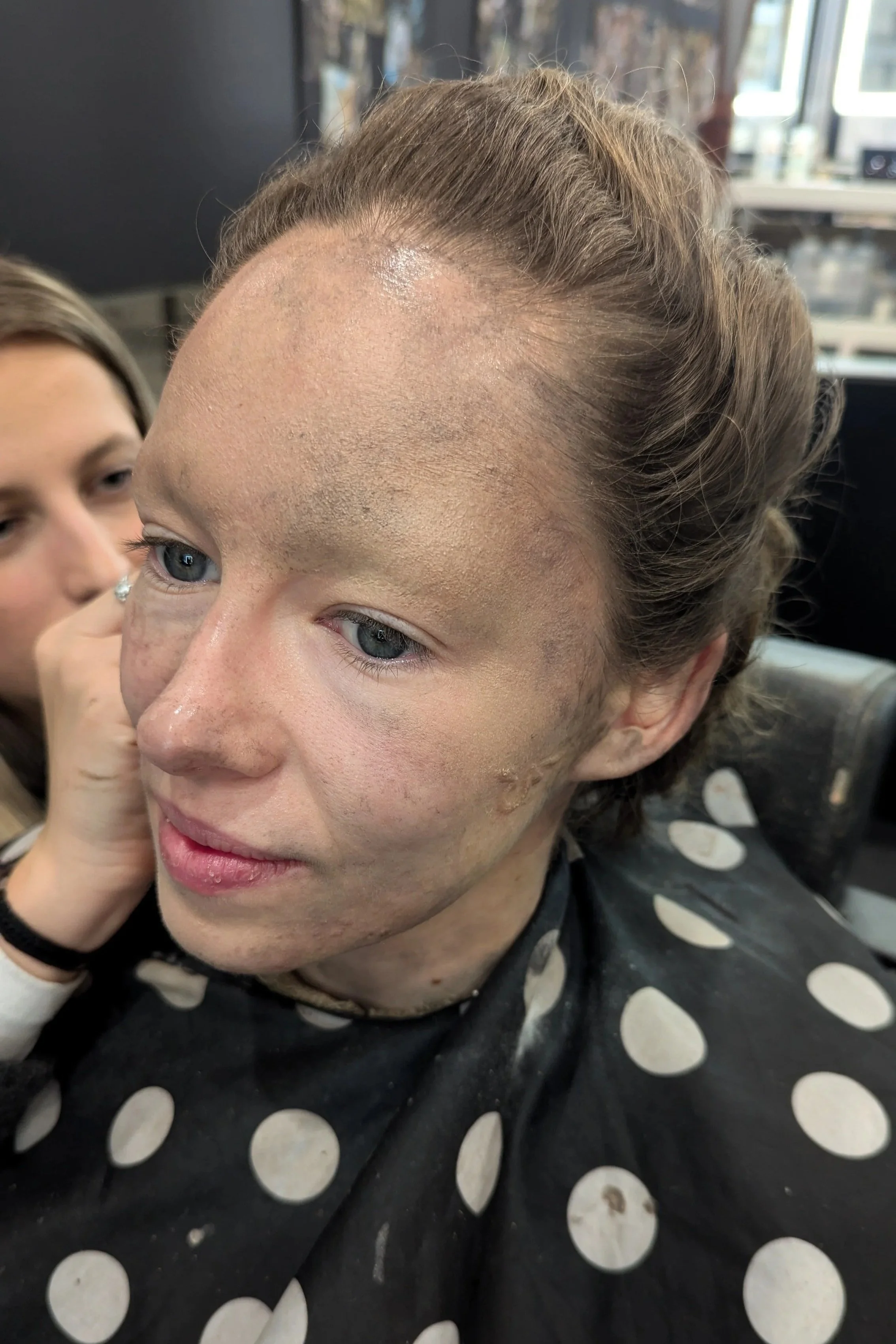 A woman with a partially shaved head and new hair growth, side profile, at a salon with a polka dot cape, and someone applying makeup or makeup powder to her face.