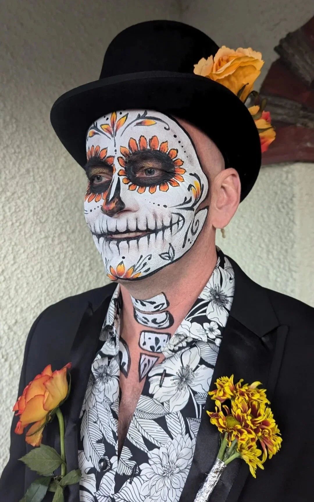 Person wearing a black hat decorated with yellow-orange flowers and a face painted in the style of a sugar skull, with black, white, and orange designs, including floral motifs and skeleton details. The person is dressed in a black floral shirt and jacket with yellow-orange flowers pinned to it.