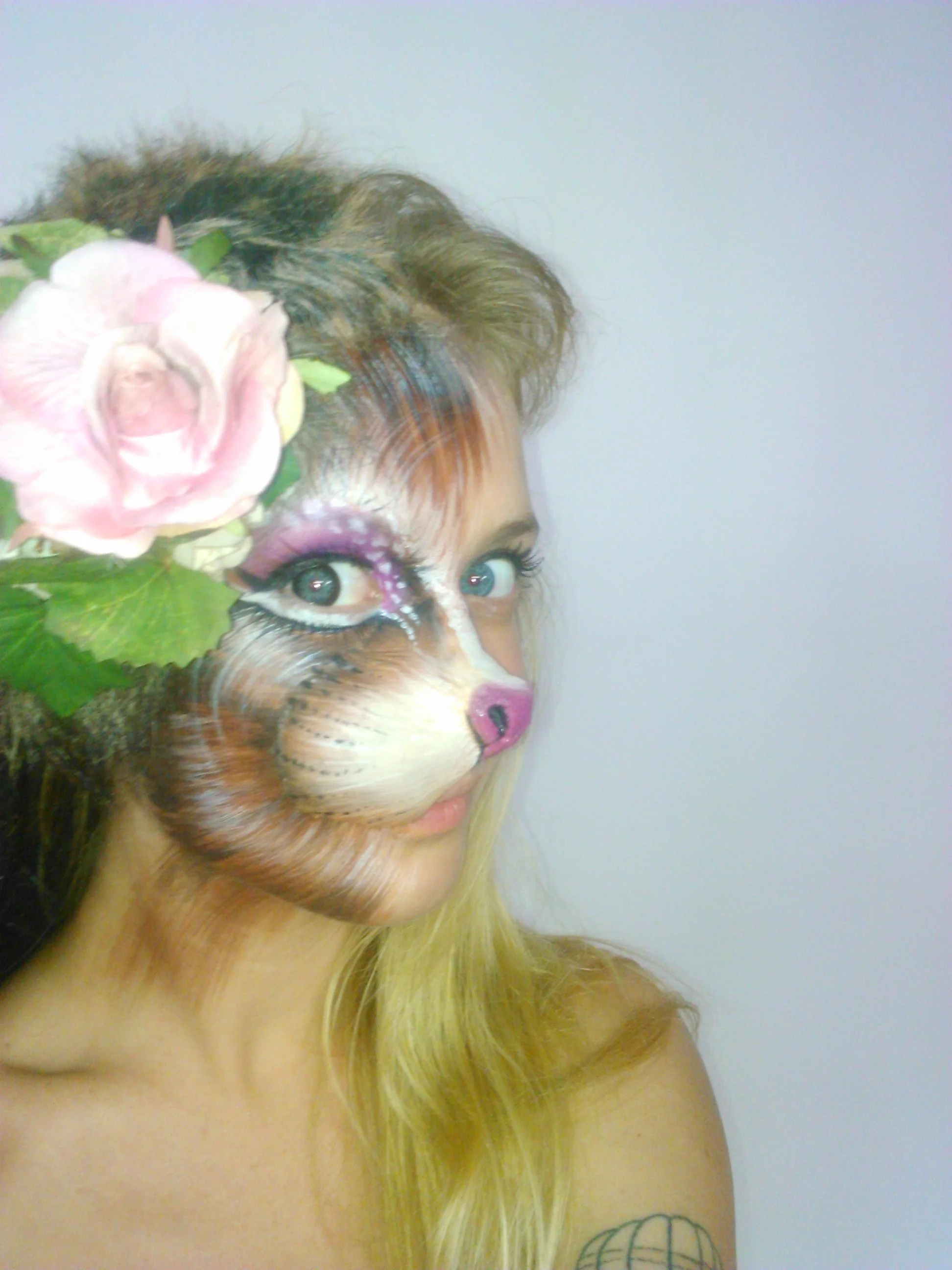 Person with face painted as a dog, large pink flower and green leaves in hair, blonde hair, tattoo on shoulder, neutral expression.