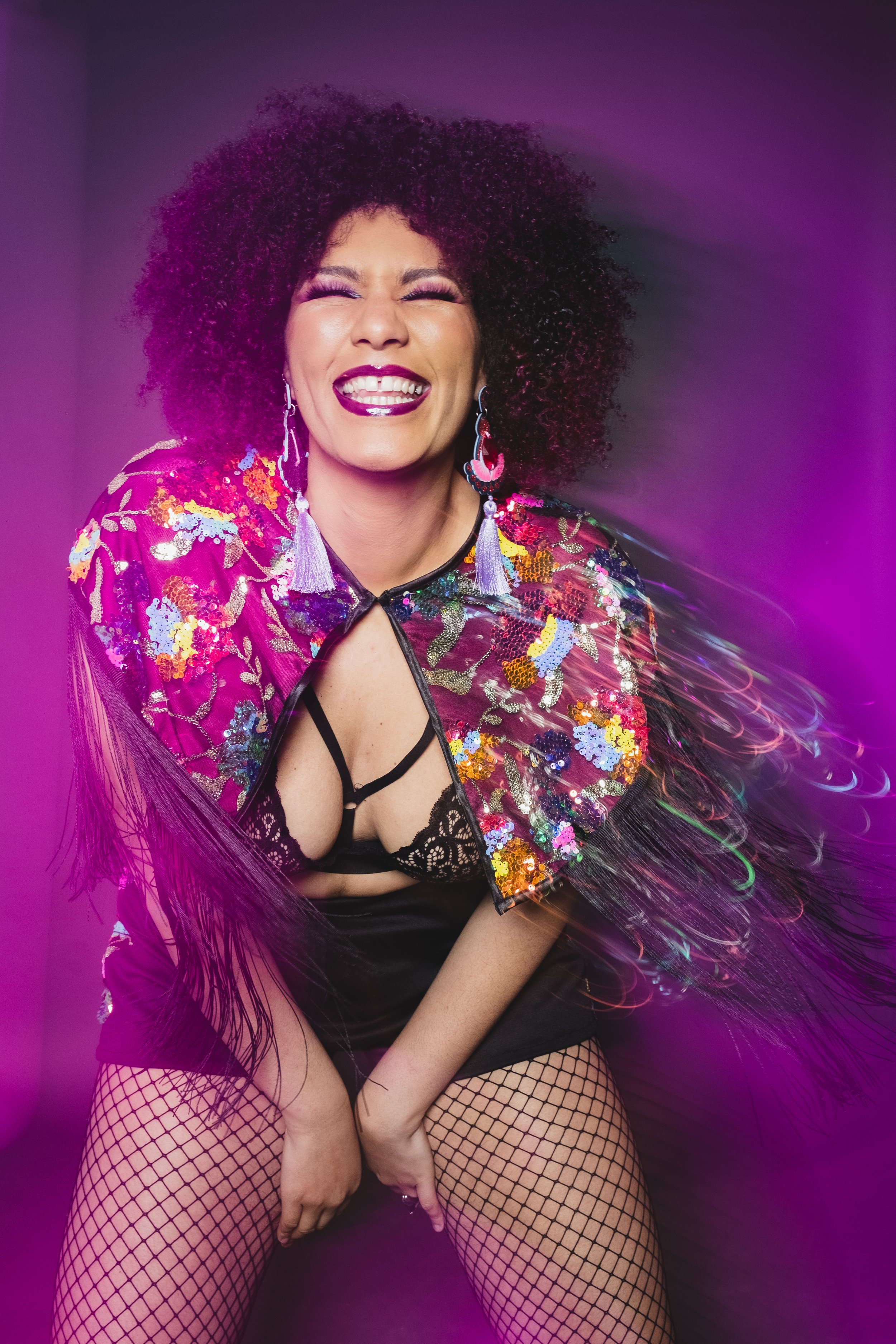 A woman with curly black hair, wearing a colorful sequin jacket, black lingerie, fishnet stockings, and statement earrings, smiling with eyes closed, pink and purple light effects.