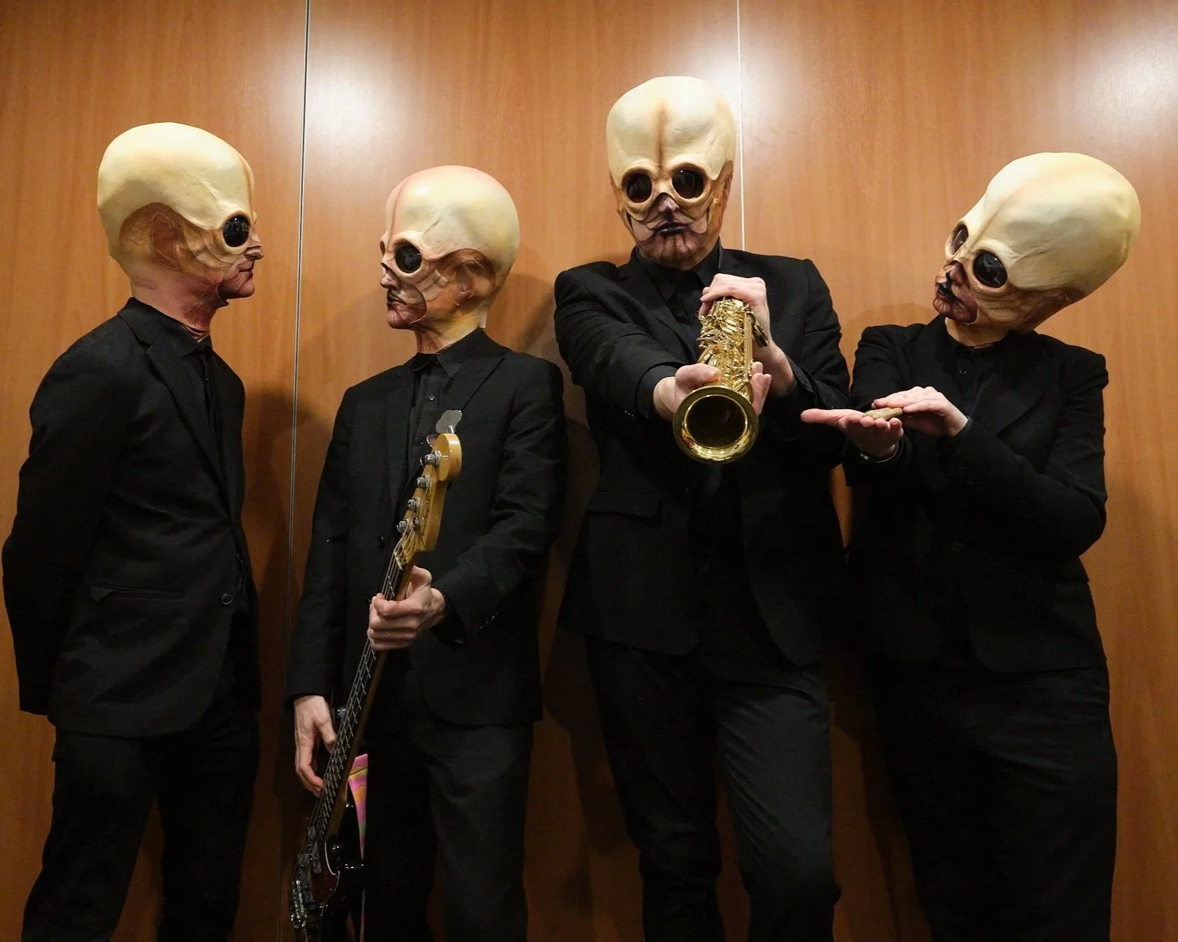 Four people wearing black suits and large silicone alien masks, with three of them holding musical instruments, standing in front of a wooden panel wall.