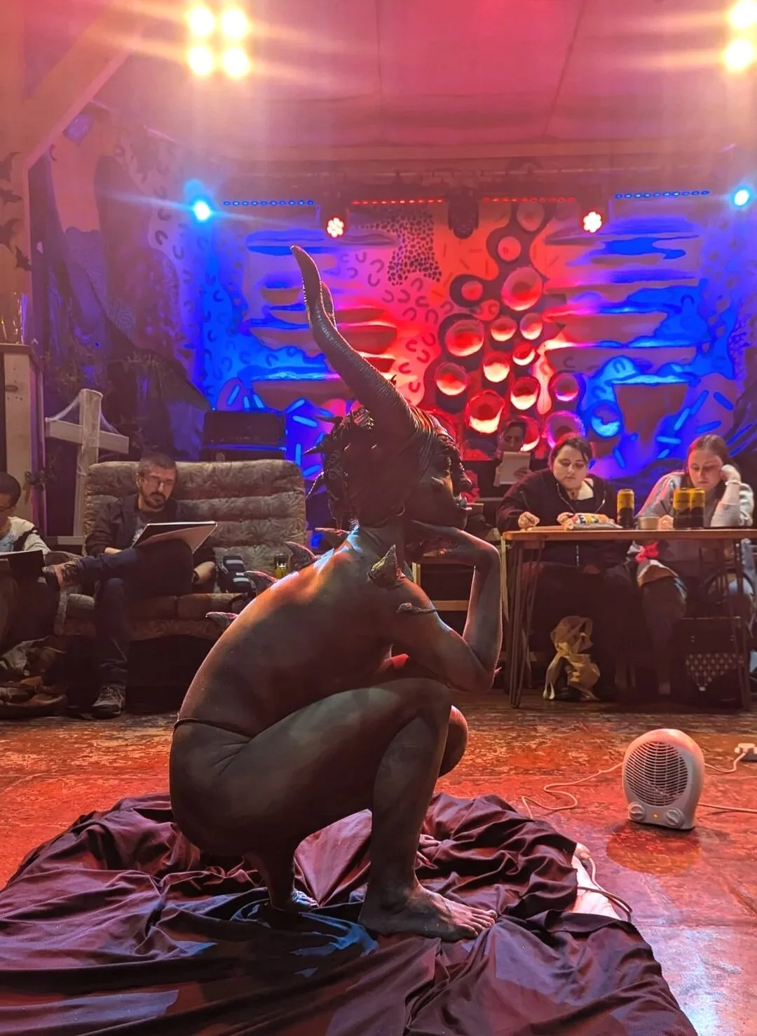 A person with a demon-like mask, including horns and a long tongue, crouches on a cloth-covered floor in a dimly lit room with colorful abstract wall art and seating. Audience members sit at tables, watching the performance, with purple and red lighting emphasizing the scene.