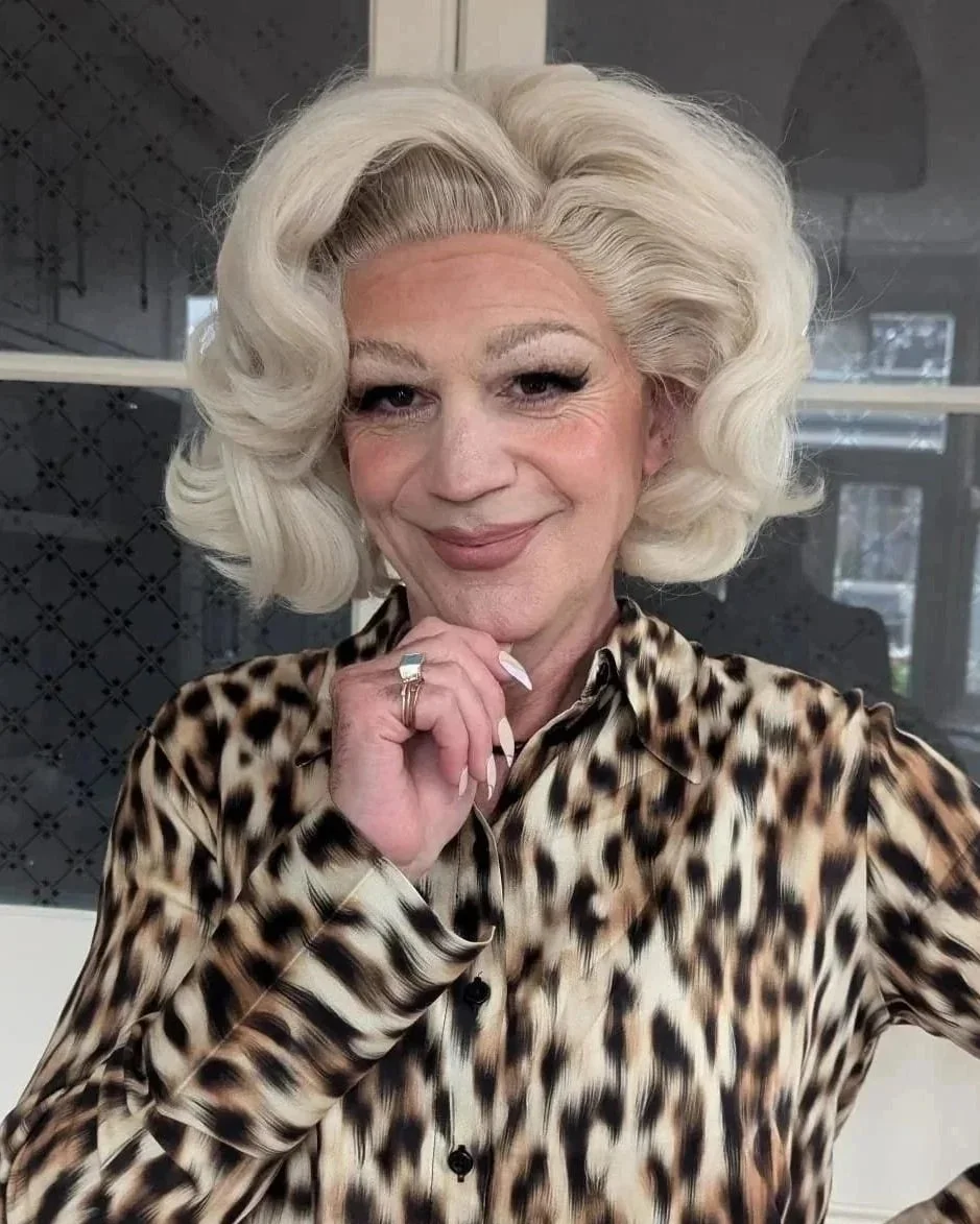 A woman with platinum blonde, styled hair, wearing a leopard print blouse, smiling, with her hand near her chin.