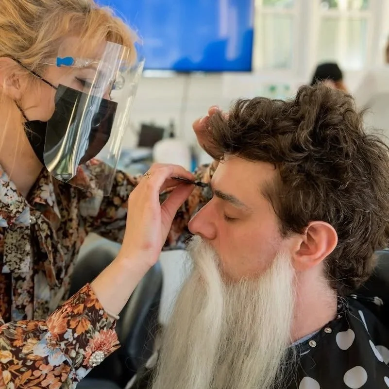 A woman with blonde hair wearing a face shield and face mask applies makeup to a man with curly hair and a long white beard.