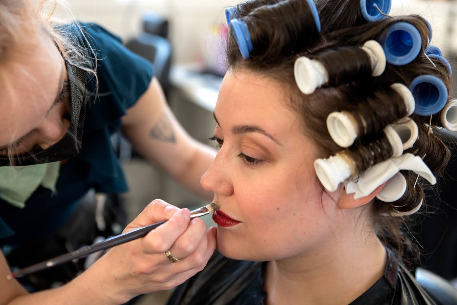 A makeup artist applies red lipstick to a woman's lips, who is seated with hair in roller curlers.