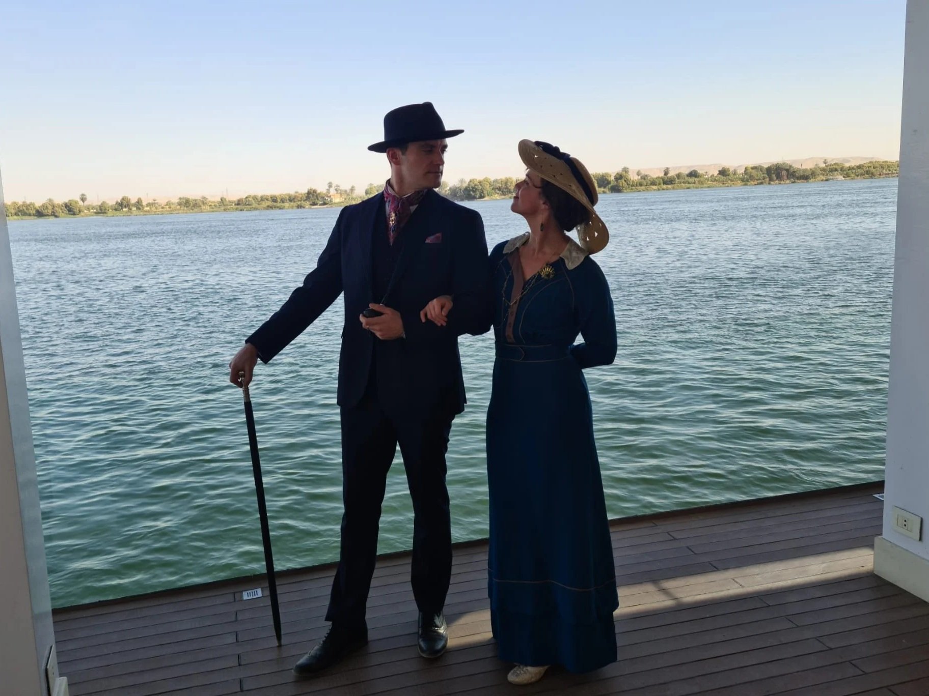 A man and woman dressed in vintage clothing standing on a wooden deck by a body of water, with a distant shoreline and trees visible in the background.