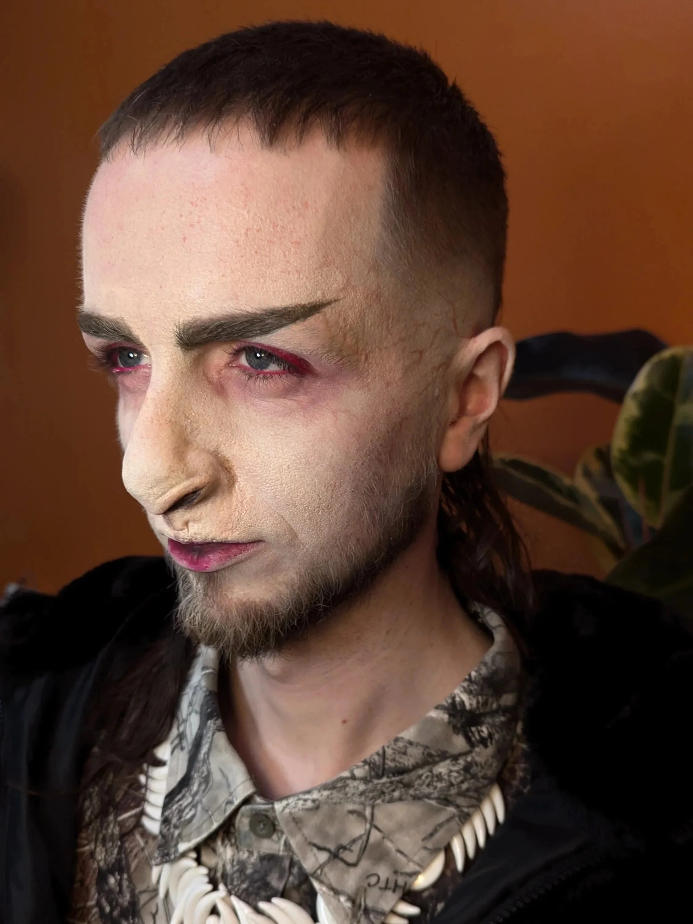 Close-up of a person with short brown hair, makeup with red accents around the eyes, light-colored face makeup, and a beard. They are wearing a camouflage shirt and a shell necklace.